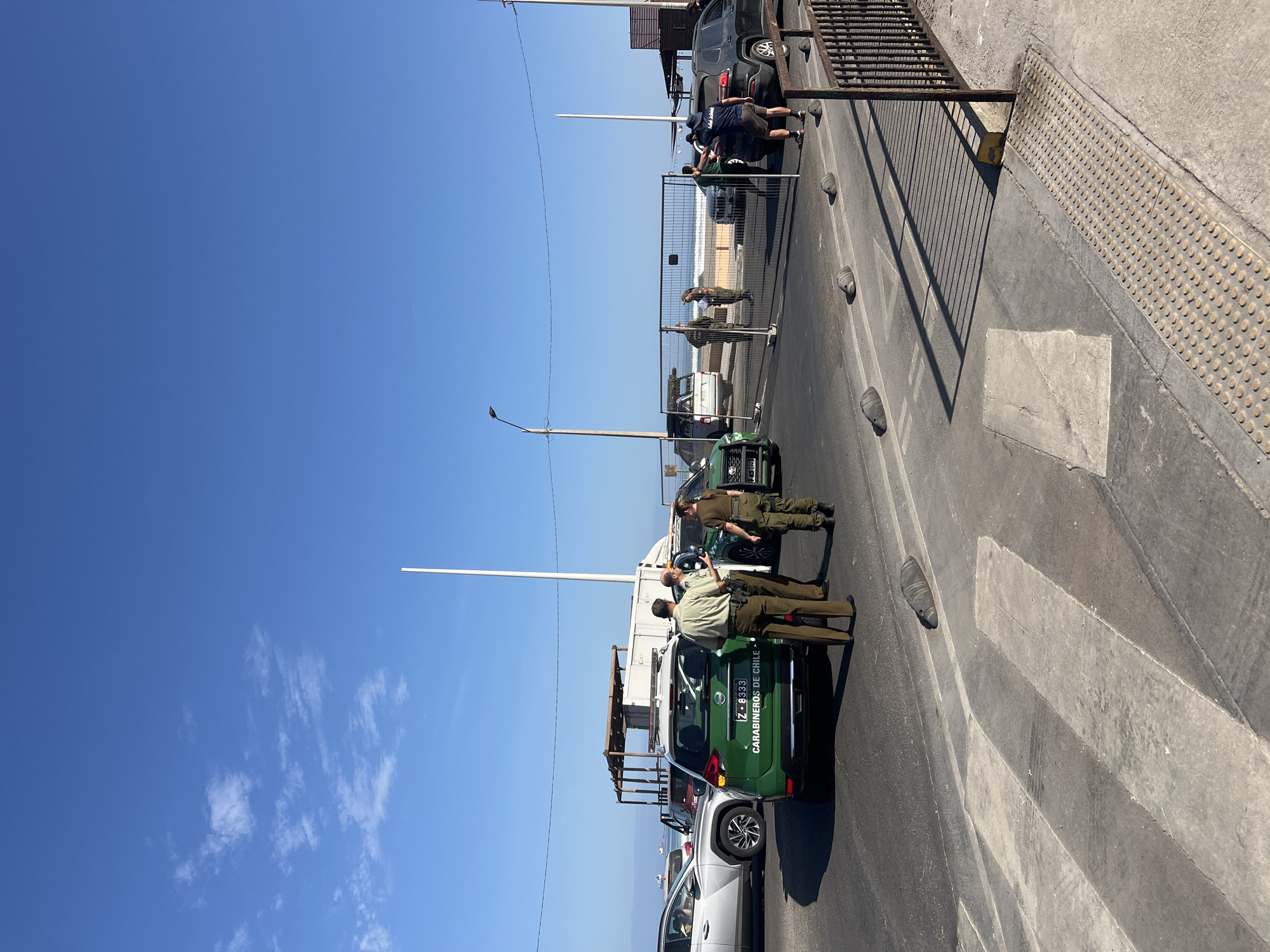 The image shows a street scene in Arica, Chile, with a focus on two Carabinero de Chile (Chilean police) officers standing next to their parked vehicle. The green and white police vehicle is parked near a crosswalk. The officers are wearing green uniforms. Several other vehicles are parked nearby, including a white car. In the background, a metal fence separates the street from an area that appears to overlook a beach or waterfront. People are seen standing near the fence. The sky is blue with a few clouds.
