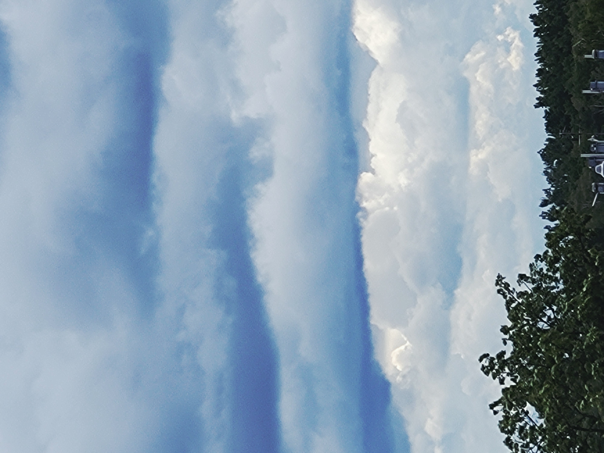 The image captures a cloudy sky over a dense line of trees. The sky is filled with layers of clouds, ranging from dark, heavy masses to lighter, fluffier formations. The clouds are a mix of blues and whites, suggesting a dynamic weather system. The top of the image is dominated by the sky, while the bottom features the upper canopy of trees, creating a stark contrast between the atmospheric and terrestrial elements. No people or specific actions are visible in the frame. The setting appears to be a natural or semi-natural area, possibly on the outskirts of a city or in a park.