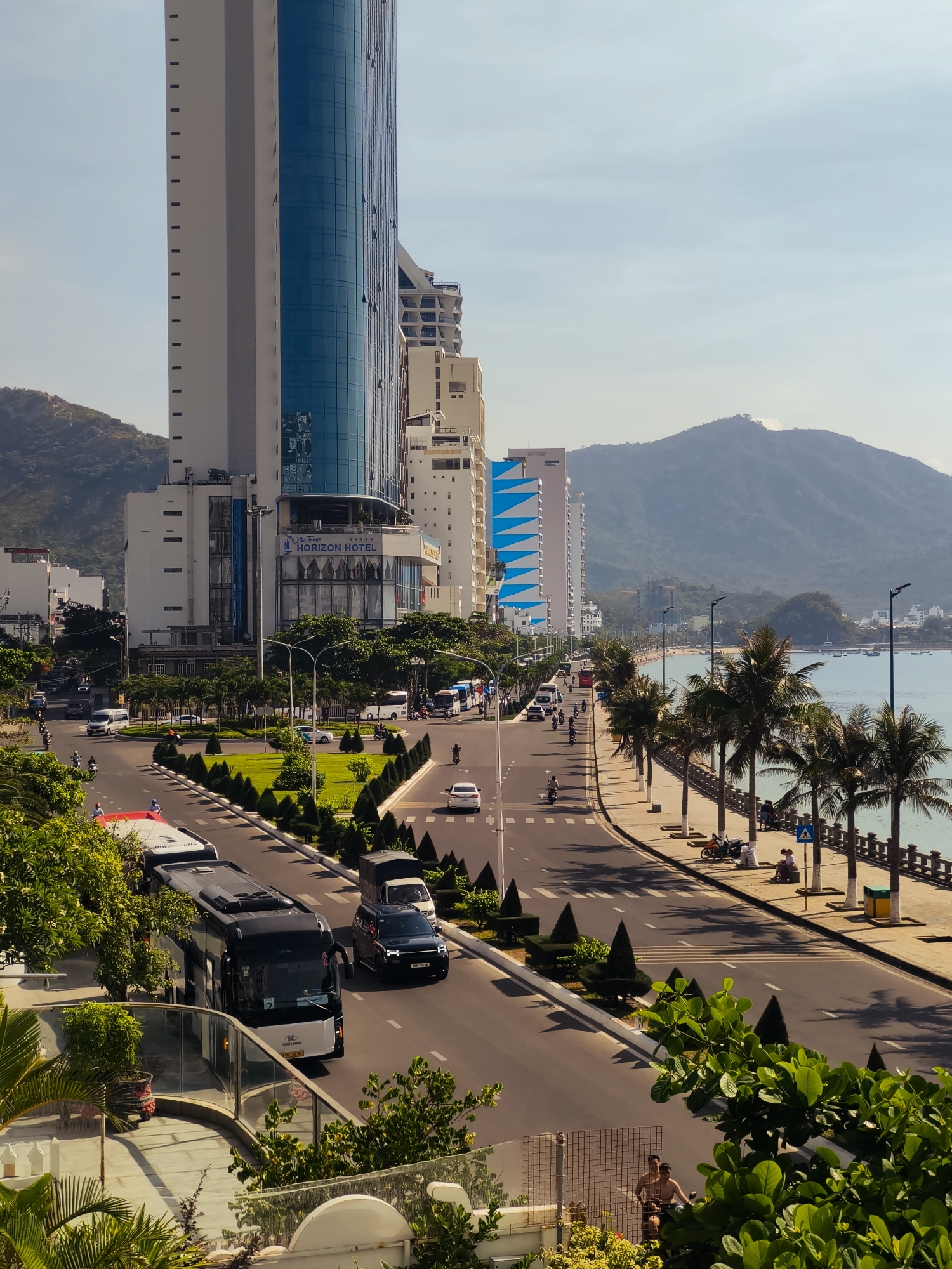 The image depicts a sunny day in Nha Trang, Vietnam, specifically along the coastal road of Bac Nha Trang Ward. A prominent, modern, tall building with a blue glass facade stands on the left side of the image, adjacent to other multi-story buildings. The road in the foreground is busy with traffic, including buses, cars, and numerous motorcycles, indicative of a lively urban environment. Lush green trees and manicured hedges line the road and a central median, contributing to the city's tropical ambiance.
Further down the road, a promenade runs alongside the beach, with palm trees offering shade. People can be seen strolling or sitting along the promenade, enjoying the coastal view. The turquoise ocean stretches out to the horizon, where a hazy mountain range provides a scenic backdrop. Boats are visible on the water, and the sky is a clear, bright blue. The overall scene captures a vibrant, coastal city in Vietnam, with a blend of modern architecture and natural beauty.