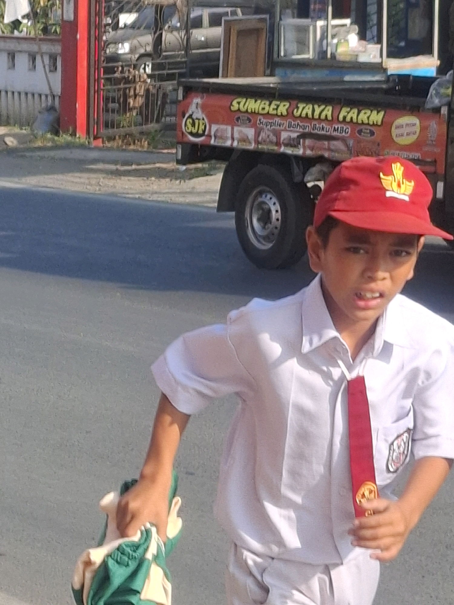 In Selajambe, Indonesia, a young boy wearing a white school uniform with a red tie and red cap walks on an asphalt road. He is holding a green and white cloth bag in his left hand. A truck with the words "SUMBER JAYA FARM" on its side is visible in the background. The truck is decorated with graphics and text advertising its services as a supplier of raw materials. The scene appears to be a typical street view with a mix of urban elements and vegetation.