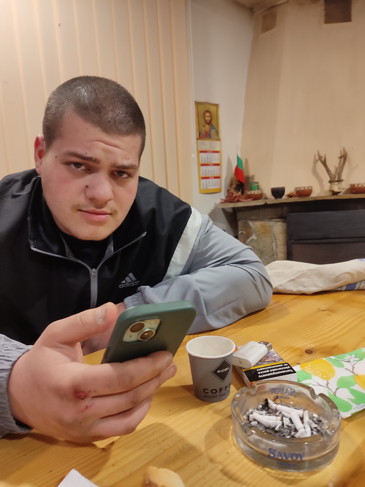 The image shows a young man sitting at a wooden table. He has short, dark hair and is wearing a black and grey Adidas jacket. He is holding a green smartphone in his left hand. On the table, next to him, are an ashtray filled with cigarette butts, a lighter, a pack of Savoy cigarettes, a paper coffee cup with "RIOBRA COFFEE" printed on it, and a napkin with a lemon pattern. A wound is visible on the back of his left hand.
Behind him, the background shows a wall with a fireplace. Above the fireplace, there are decorative objects, including deer antlers. To the left of the fireplace, a calendar with an icon of Jesus and the Bulgarian flag is hanging on the wall. The location is likely in the city of Varshilo, Bulgaria.