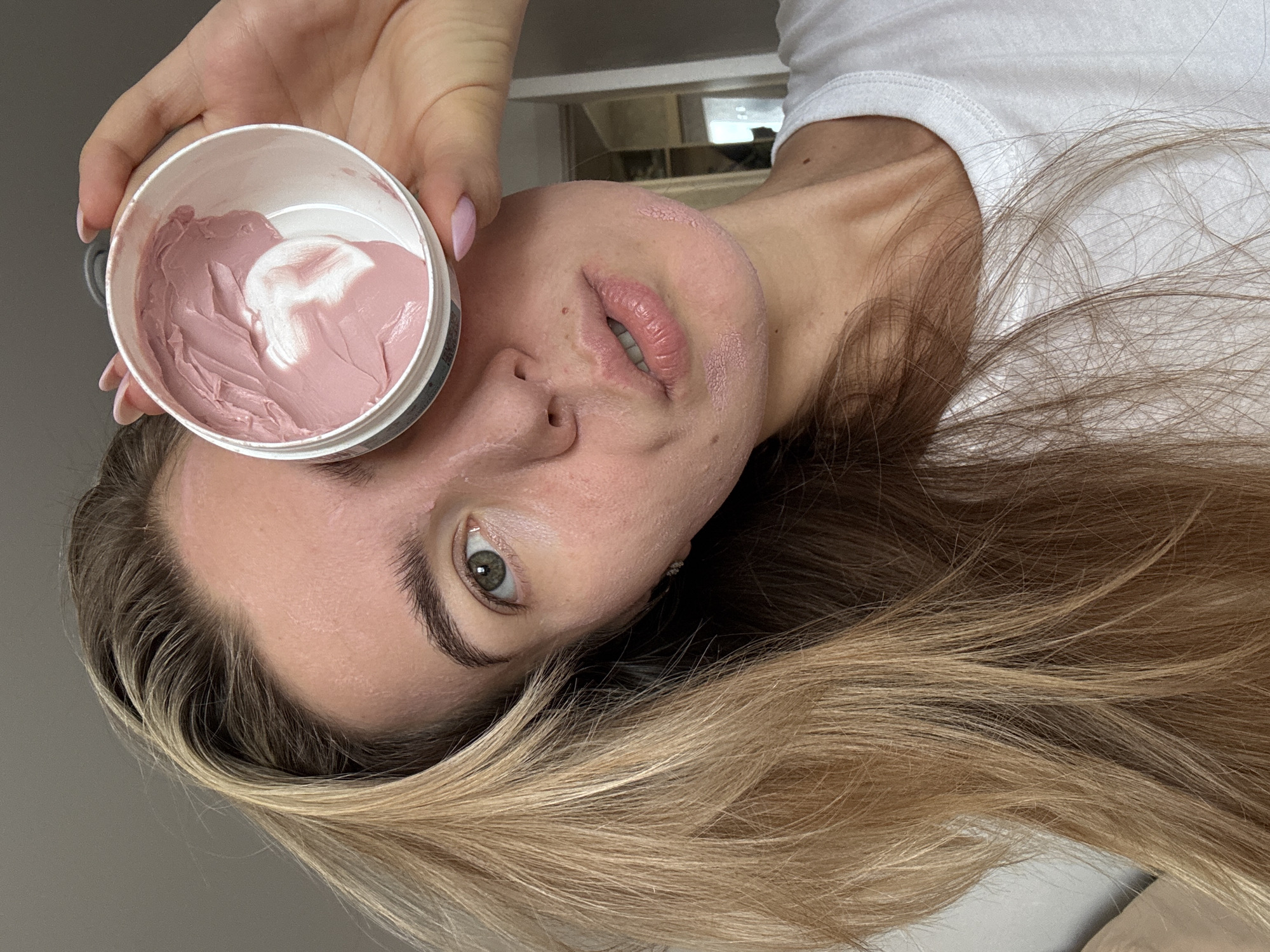 A close-up, selfie-style shot shows a young Caucasian woman with blonde hair holding a small, open jar of pink clay mask in her right hand, close to her right eye. She is wearing a white t-shirt. The woman's face is partially covered by the jar. Her skin appears dry and flaky in some areas, particularly on her chin and jawline. Her lips are slightly parted and appear dry. A smudge of the pink mask is visible on her right cheekbone and under her chin. Her eyebrows are well-defined. The background is out of focus, showing a neutral wall and a hint of what appears to be an interior space with a doorway.