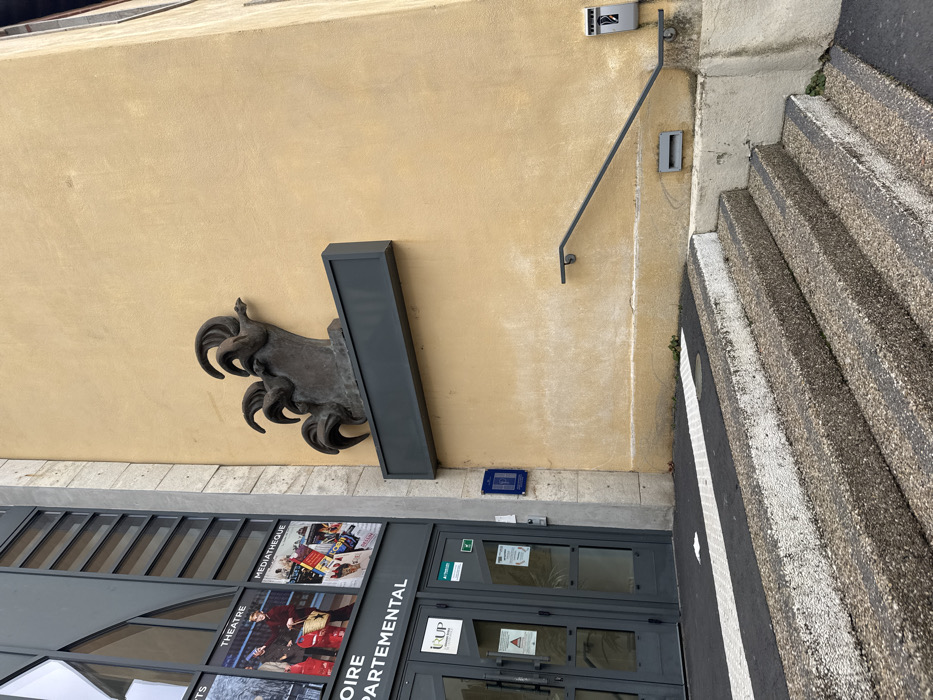 The image depicts the exterior of a public or institutional building in Le Puy-en-Velay, France, specifically associated with a "Médiathèque" and "Départemental" services, indicated by banners on the facade, and an "IURP" logo on the entrance doors.
The scene is dominated by two distinct architectural styles: a modern, dark grey panelled facade with large glass windows on the left, and an older, light yellow stucco wall on the right, exhibiting some weathering and discoloration. The main entrance, located on the left facade, consists of double grey-framed glass doors. Above these doors are banners advertising "THEATRE", "MÉDIATHÈQUE", and "HISTOIRE DÉPARTEMENTAL".
A prominent dark-colored, possibly bronze, abstract sculpture of three stylized roosters (coqs) is mounted on the yellow stucco wall, positioned above a dark, empty rectangular plaque. The roosters feature flowing, curvilinear forms suggesting their combs and tail feathers.
Access to the building is via a set of exposed aggregate concrete stairs with dark treads and lighter risers, leading upwards from the foreground. The top surface of these steps is marked with black and white stripes. A black metal handrail is affixed to the yellow wall alongside the right side of the stairs. Below the handrail, a small grey rectangular vent or grate is visible at ground level. Further right, a grey wall-mounted utility box or service panel is present. A blue informational or historical plaque is affixed to the wall near the double doors. The immediate ground in the foreground consists of dark pavement with a white striped section.
No individuals are present in the scene, and no explicit actions or interactions are depicted. The lighting is even, suggesting an overcast day.