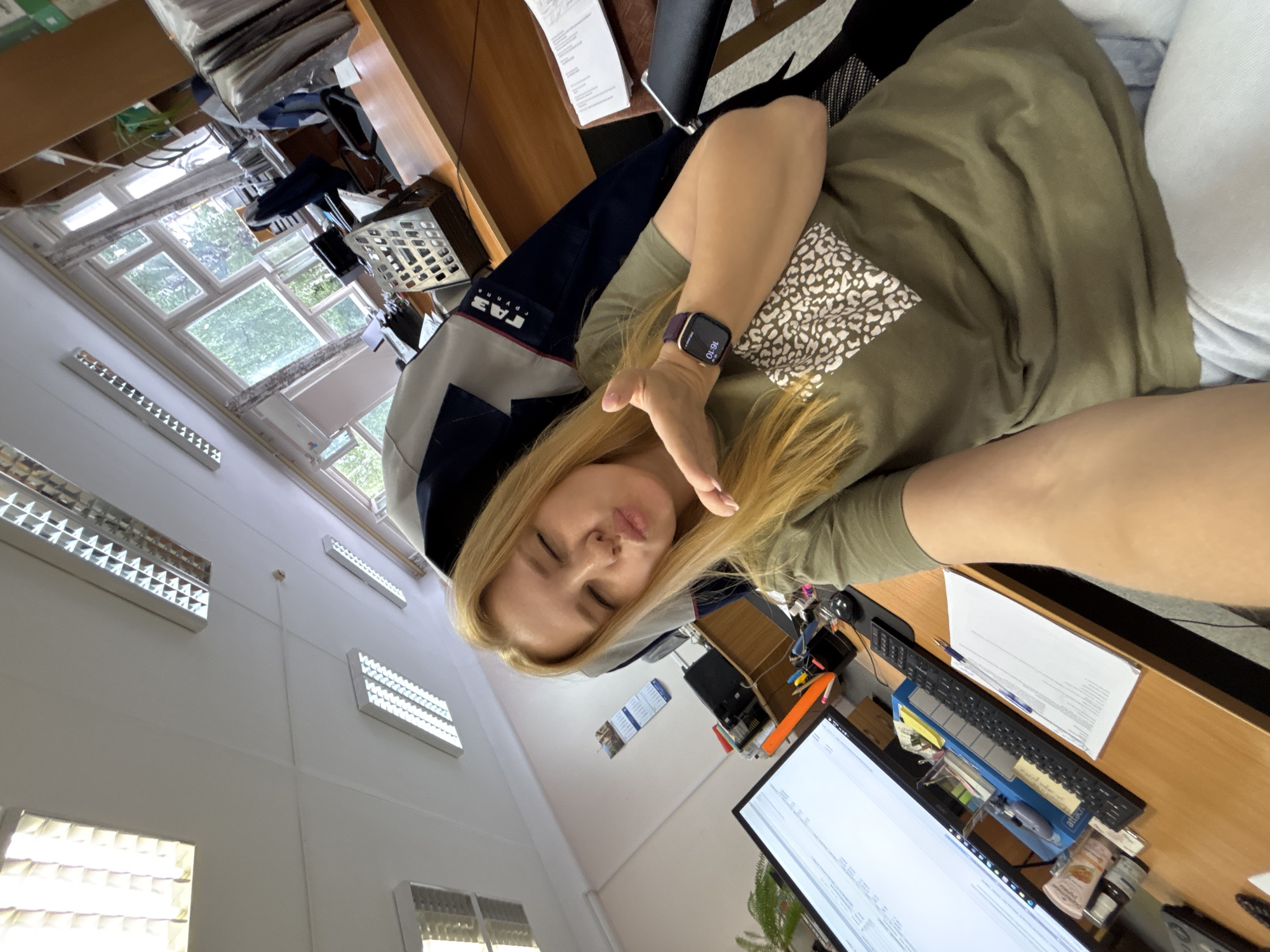 A blonde woman wearing a green t-shirt and light blue pants is taking a selfie in an office in Zavolzhye, Russia. She is sitting in an office chair, with her right hand raised and her lips puckered as if blowing a kiss. She is wearing a dark-colored smartwatch on her left wrist. The office is equipped with fluorescent lighting, windows with greenery visible outside, and wooden desks with office supplies, including a computer monitor, keyboard, pens, and papers. A blue chair with a white and blue emblem is behind her.