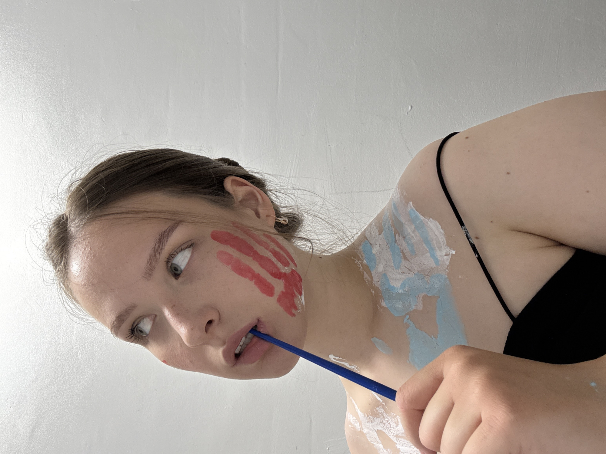 The image shows a young woman with light skin, light brown hair pulled back, and green eyes looking upwards and to her left. She has red paint applied as handprints on her cheek and chest. She is also wearing light blue and white paint on her chest and shoulder. Her lips are slightly parted as she holds a blue paintbrush in her mouth. She is wearing a black tank top. The background is a plain white wall. There is no visible indication of her location in Chaikovsky, Russia.