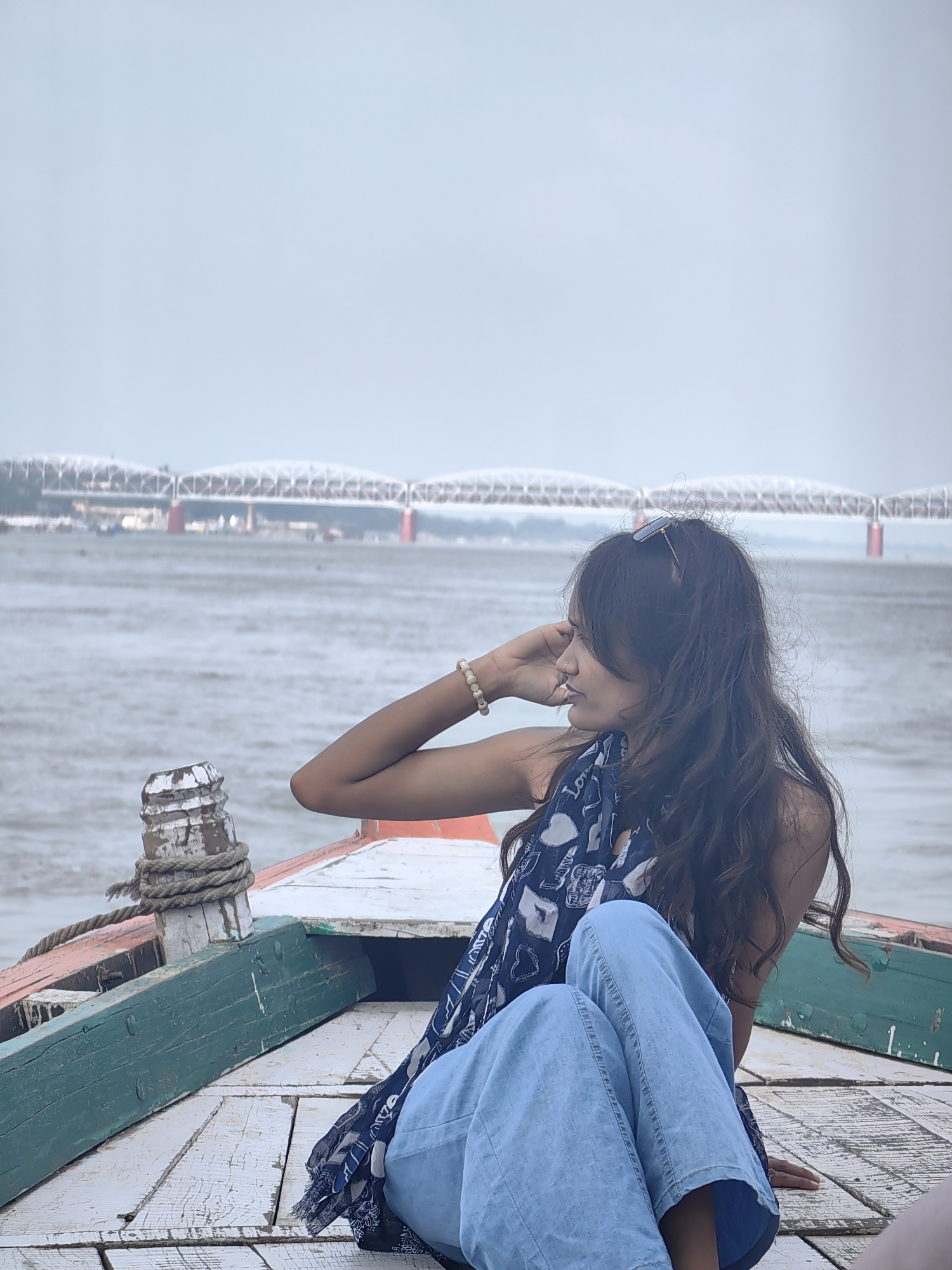 A young woman sits on the wooden deck of a boat on the Ganges River in Varanasi, India. She is wearing a blue and white patterned scarf and light blue wide-leg pants. Her hair is long and dark, and she is looking away from the camera with her hand resting on her forehead. Sunglasses are perched on top of her head. In the background, a large, red-pillared bridge spans the river under a cloudy sky. The boat's wooden planks are weathered and painted in shades of white, green, and orange, with a thick rope tied to a bollard in the foreground. The river appears murky and choppy.