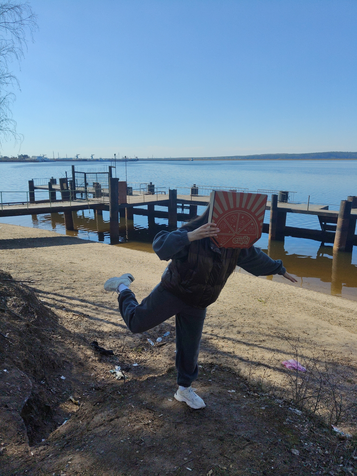 A person stands on the sandy shore of a body of water, holding a pizza box in front of their face. The person is wearing a grey sweatsuit and white sneakers, and they are striking a pose with one leg raised and arms extended. Behind the person is a pier with wooden pilings and a metal railing. In the background is a large body of water and the coastline of what appears to be the city of Unknown, Unknown. The sky is clear and blue. The foreground shows patches of grass and debris littering the ground.