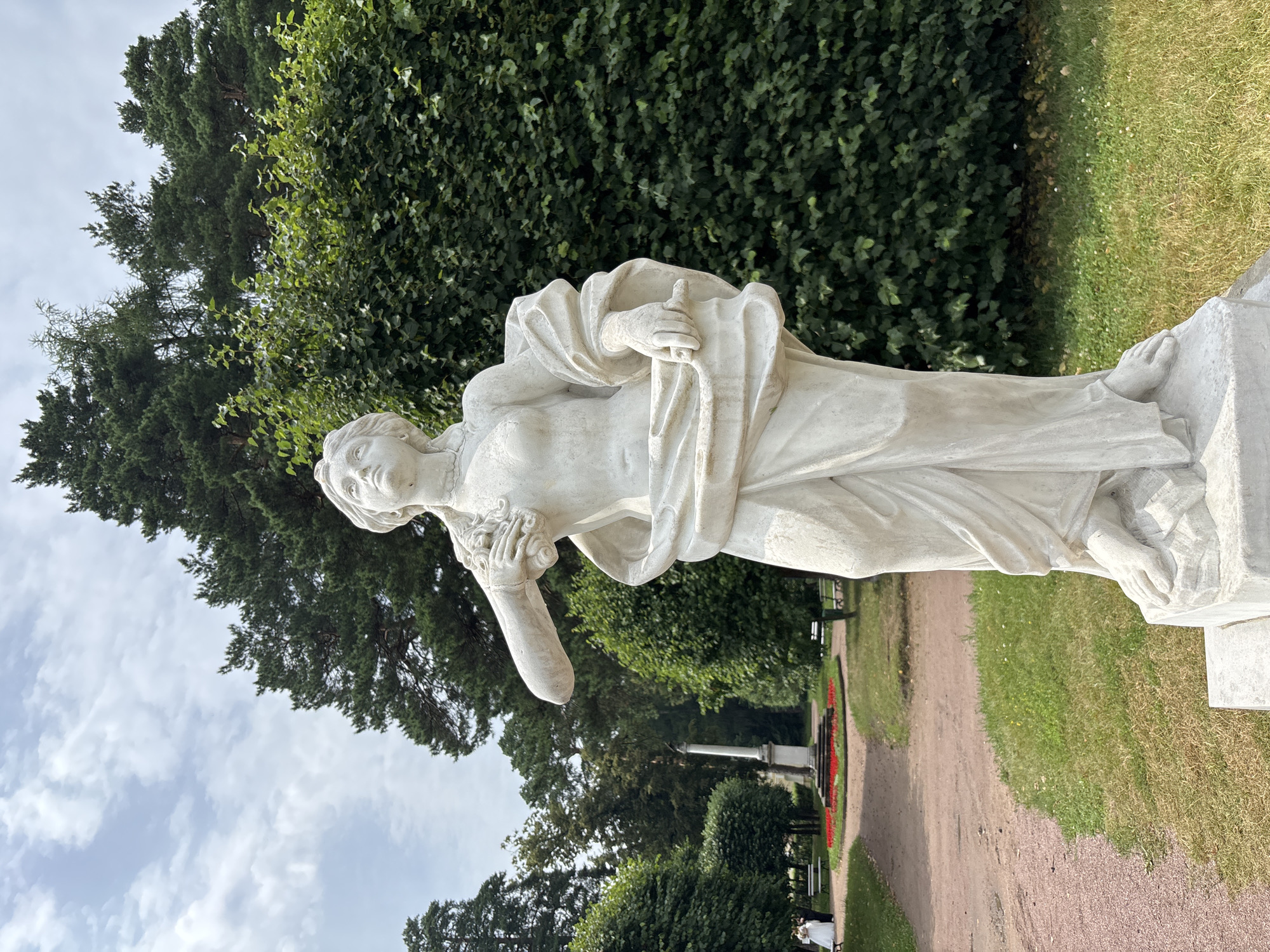 An outdoor, eye-level, full-shot photograph depicts a white marble sculpture of a woman in the foreground, set against a backdrop of lush green trees and a partly cloudy sky. The sculpture, positioned centrally, is of a female figure with draped clothing and flowing hair. Her left arm is raised, holding what appears to be flowers or grapes. Her right hand is positioned near her waist, holding a fold of her garment. The sculpture stands on a pedestal in a park-like setting.
In the middle ground, a pathway curves through the grass, leading towards a monument with a column. Several benches are visible along the path. On the left side of the pathway, two figures, a bride in a white dress and a man in dark attire, are standing and facing away from the camera. A bed of red flowers is planted in a circular pattern around the base of the monument.
The background is dominated by tall, dense green trees and a patch of blue sky with scattered white clouds. The overall environment suggests a well-maintained park or garden, characteristic of the Arkhangel'skoe Estate, a suburban cultural complex and former country estate of the Yusupov family, located in Krasnogorsky District, Moscow Oblast, Russia, which is often associated with Arkhangel'skoe, a town in Arkhangelsk Oblast. The lighting indicates it is daytime, with soft sunlight filtering through the clouds.