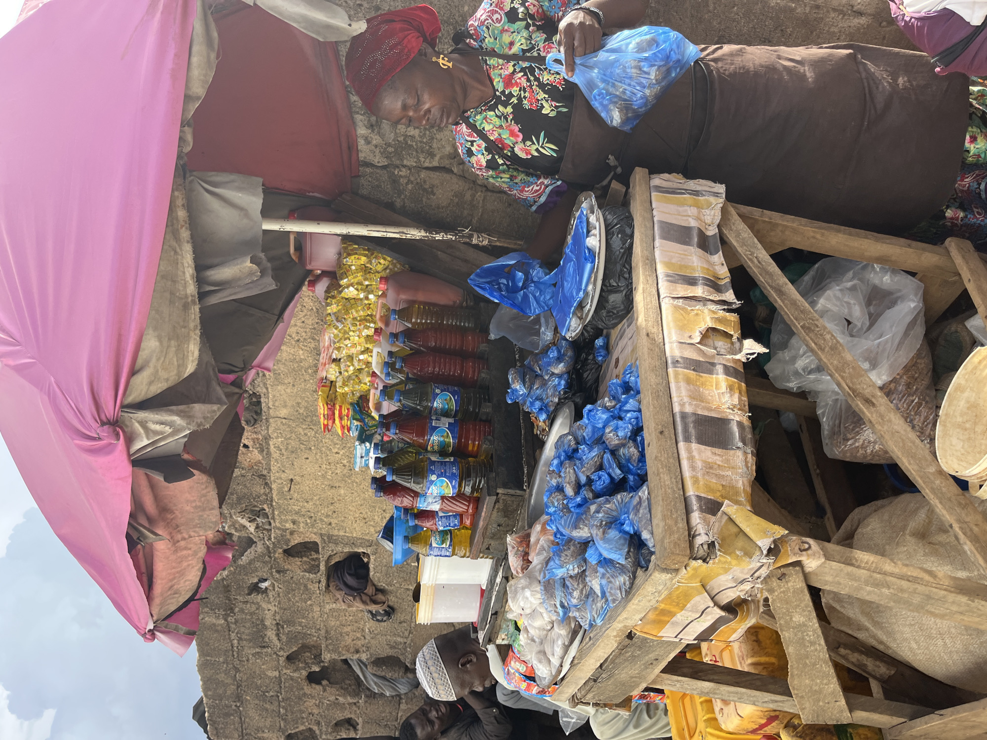 The image depicts an outdoor market scene in Rafin Par, Nigeria. In the foreground, a woman wearing a red headscarf and a floral print shirt over a brown apron is standing behind a wooden stall. She is holding a blue plastic bag filled with goods. The stall is laden with various items, including numerous small blue plastic bags containing what appear to be dried fish or similar food items, as well as white plastic bags of produce. Behind the stall, on a shelf, are rows of bottled liquids, including what looks like cooking oil in yellow bottles and other beverages in red and blue bottles. To the left of the stall, two men are visible. One is wearing a white cap and a patterned shirt, seated and looking down. Another man, partially obscured, is seated behind him. The backdrop consists of a rough, stone wall with irregular openings. A large, pink, tattered umbrella shades the stall and its contents. The overall environment suggests a bustling, informal market setting.