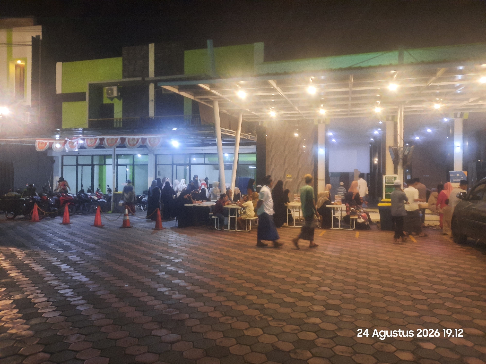 The image depicts an outdoor scene at night, with a group of people gathered in what appears to be a public space with a covered walkway. The ground is paved with hexagonal interlocking bricks. In the background, there is a building with green and black architectural elements and white lights illuminating the covered area.
Several individuals are visible, many of whom are wearing traditional Islamic attire, including hijabs and long robes. Some are seated at tables, suggesting a meal or meeting is taking place. Others are standing and interacting. To the left, a few motorcycles are parked, and orange traffic cones are placed on the ground. A black car is partially visible on the right side of the image.
The lighting suggests an artificial environment, likely from overhead lamps and lights from the building. The overall atmosphere appears to be one of a community gathering or event. A date stamp in the lower right corner reads "24 Agustus 2026 19.12".