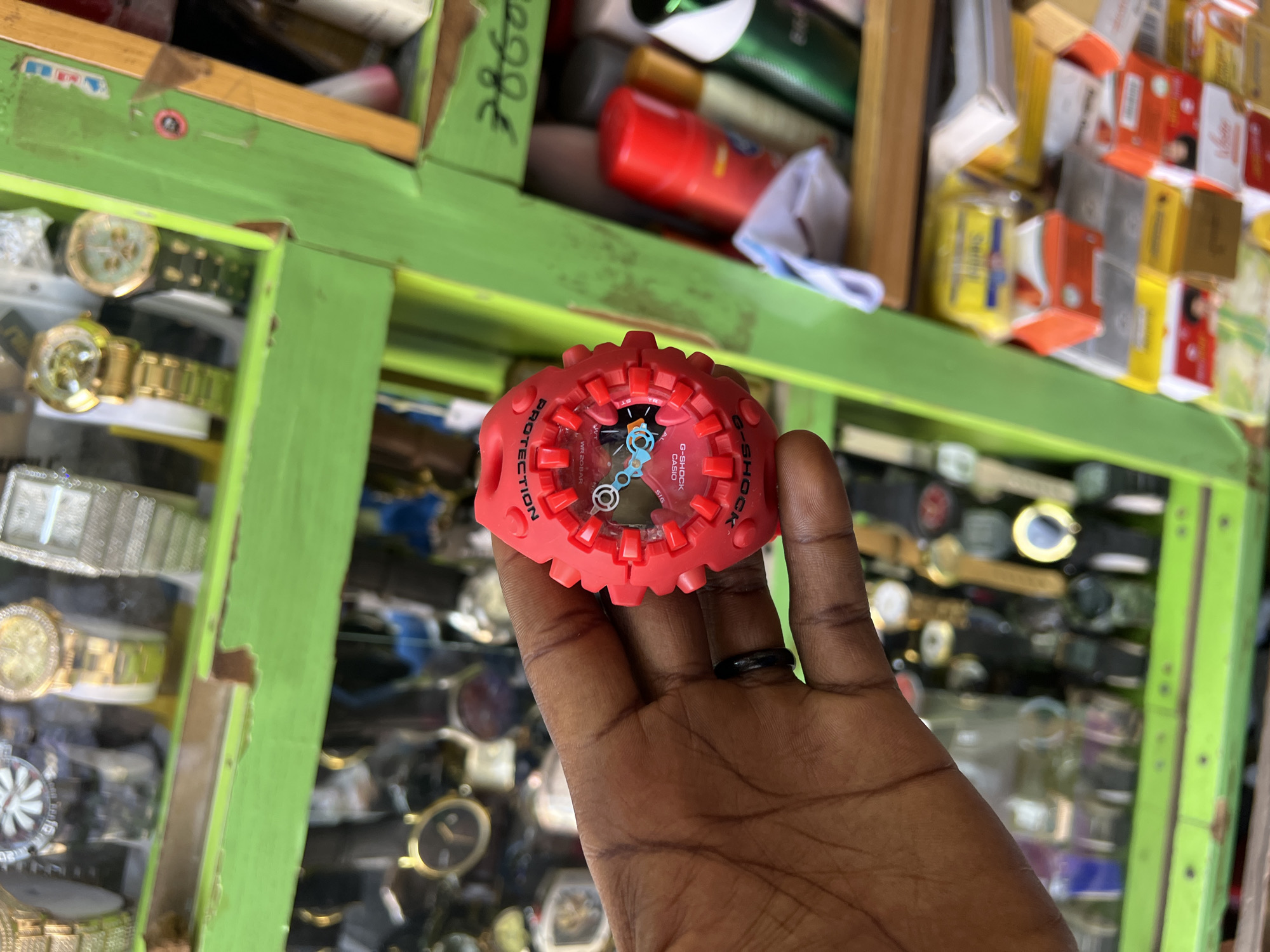 This is a medium shot, eye-level, close-up photograph of a person's left hand holding a red G-Shock watch in front of a display of watches and other items. The background is a blur of various watches in different styles and colors, including gold, silver, and black. The watches are arranged on green shelves, with some items, possibly cosmetics or toiletries, visible on a shelf to the right. The overall environment appears to be a market or street vendor stall, suggesting a commercial setting. The image was taken in Bukuru, Nigeria.
