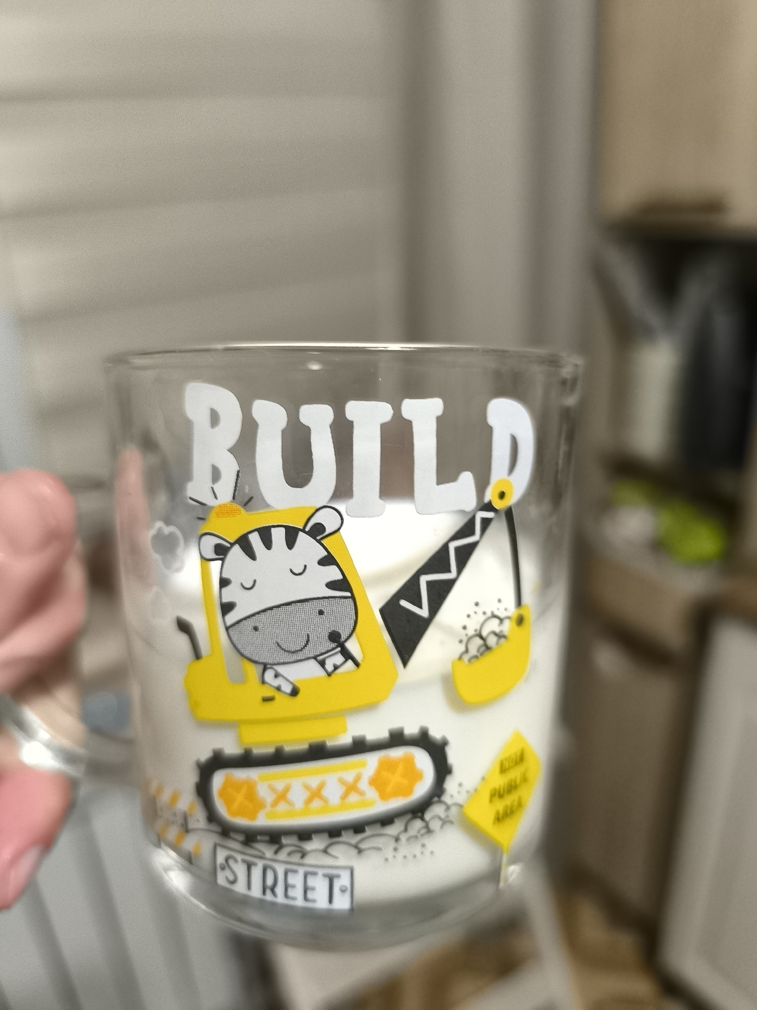 The image shows a clear glass mug with a white candle inside. The mug has a cartoon decal on it, featuring a yellow construction vehicle operated by a zebra character. The word "BUILD" is written in large white letters above the vehicle. The zebra is wearing a construction helmet and has a smiling expression. The construction vehicle has a bucket that appears to be filled with some white material, possibly the wax for the candle. The mug also has other small graphic elements, including what looks like a road sign that says "PUBLIC AREA" and a license plate that says "STREET". The background of the image is blurred, but it appears to be an indoor setting, possibly a kitchen or living area, with cabinets and a countertop visible. The lighting suggests it is daytime. There is no discernible connection to the city of Horishni Plavni, Ukraine, in the image itself.