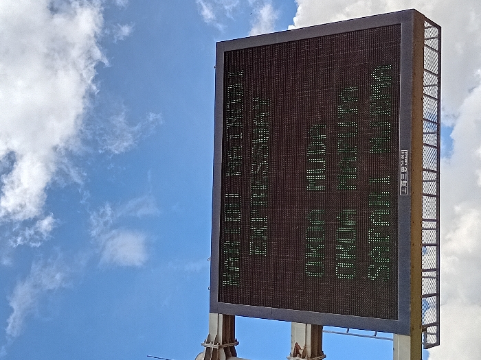 The image shows a large digital sign mounted on a metal frame, located in Mlolongo, Kenya, at latitude -1.401, longitude 36.944. The sign is made of black metal with a dark grey face that displays green text. The sign displays the following text:
"Karibu Welcome
Enthusiastic
Good Morning
Good Afternoon
Good Evening"
The sign is set against a bright blue sky with white clouds in the background. The sky is mostly clear. The sign is the only subject in the image. The sign is located on the side of a road, likely near a major thoroughfare.