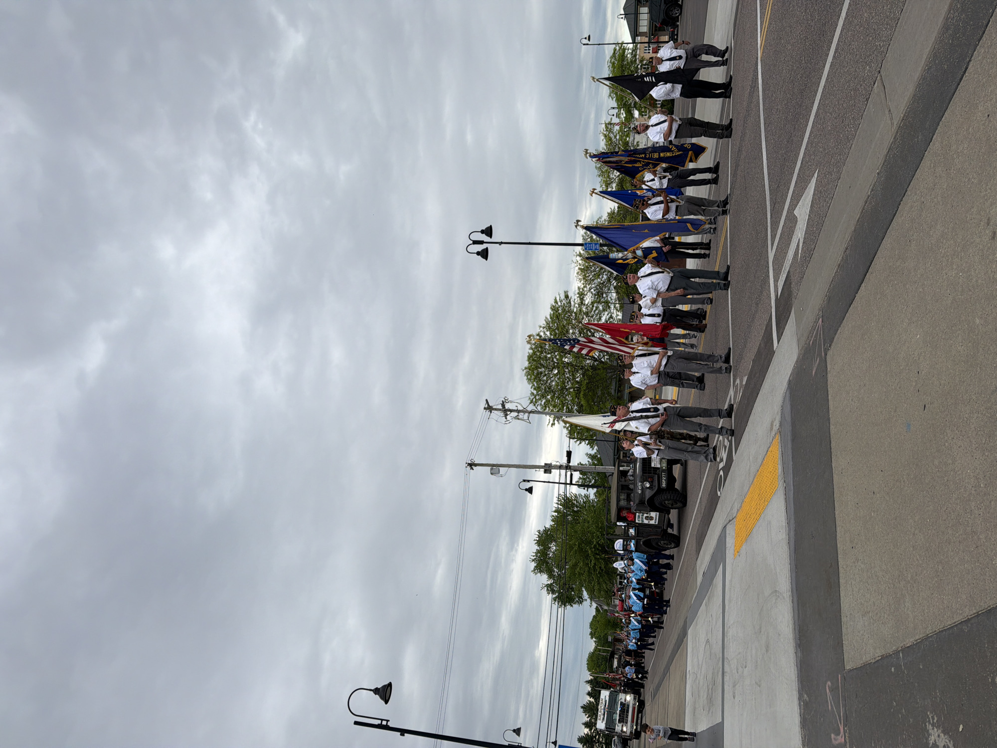 The image captures a parade in Wisconsin Dells, United States of America. A color guard with flags, including the American flag, and other participants, are seen marching along a street lined with streetlights. The street has lane markers and crosswalks. A military vehicle is parked on the side of the road. Overcast skies dominate the background.