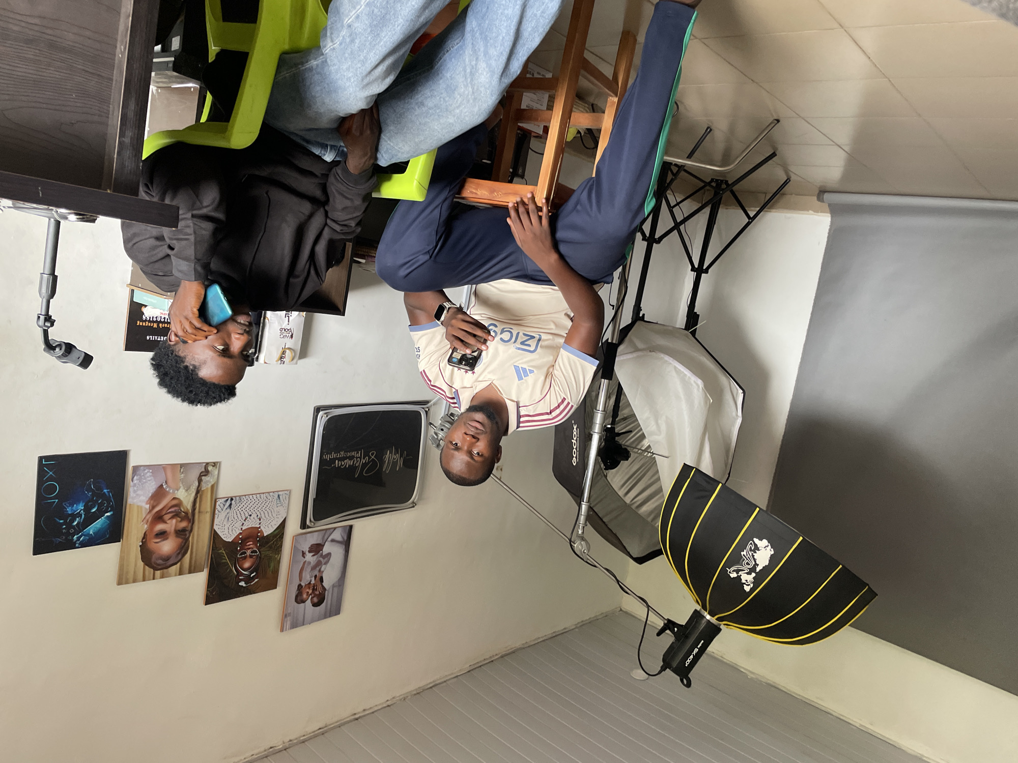 The image depicts an indoor studio setting, possibly in Jos, Nigeria, judging by the presence of numerous framed portraits on the wall. Two individuals are present. On the left, a man in a cream-colored athletic jersey with blue and green accents sits on a wooden stool, holding a smartphone. He has dark skin and is looking towards the camera. To his right, another man with dark skin is seated on a lime green chair, engaged in a phone conversation, holding a turquoise smartphone to his ear. He is wearing a black hooded sweatshirt and blue jeans.
The studio is equipped with professional photography gear. A large softbox light with yellow trim is prominently featured on the left, angled downwards. Behind it, another lighting setup with a white diffusion panel is visible. A grey backdrop is partially visible on the left side of the frame. The walls are painted a light color, and several framed pictures are displayed on the right wall. These include wedding photos, portraits of women, and a graphic print. A dark wooden desk with some items on it, including what appears to be a microphone stand, is positioned on the right. The floor is tiled in a light beige color. The overall impression is that of a functional photography studio space.
