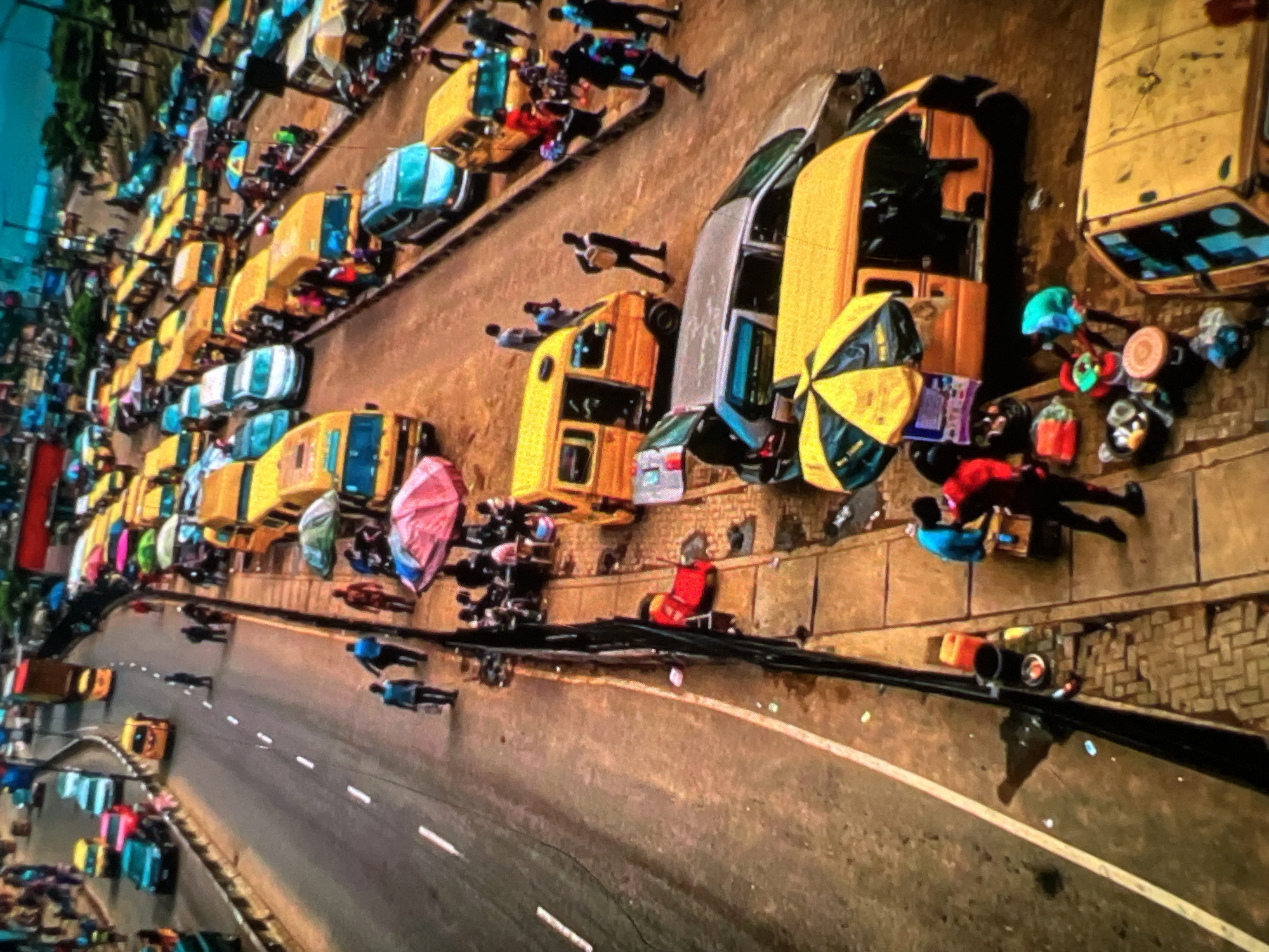 This is a high-angle, full shot of a busy street in Lagos, Nigeria, likely during the day, showing a traffic jam. The street is a multi-lane highway with a high volume of yellow minibusses, commonly known as "danfo" buses, which are a hallmark of Lagosian public transport. Several cars and a large truck are also visible, contributing to the congestion.
People are seen walking along the sidewalk and in the street, some appearing to be passengers waiting for transportation or vendors selling goods. One person is seen lying on the ground next to a red object on the sidewalk. Vendors have set up stalls under umbrellas, indicating commercial activity along the roadside. The environment is characterized by the extensive use of yellow vehicles and the bustling, crowded atmosphere typical of Lagos. The road surface appears worn, and there is litter scattered along the sides of the road and sidewalk. The overall scene depicts a chaotic but vibrant urban environment.