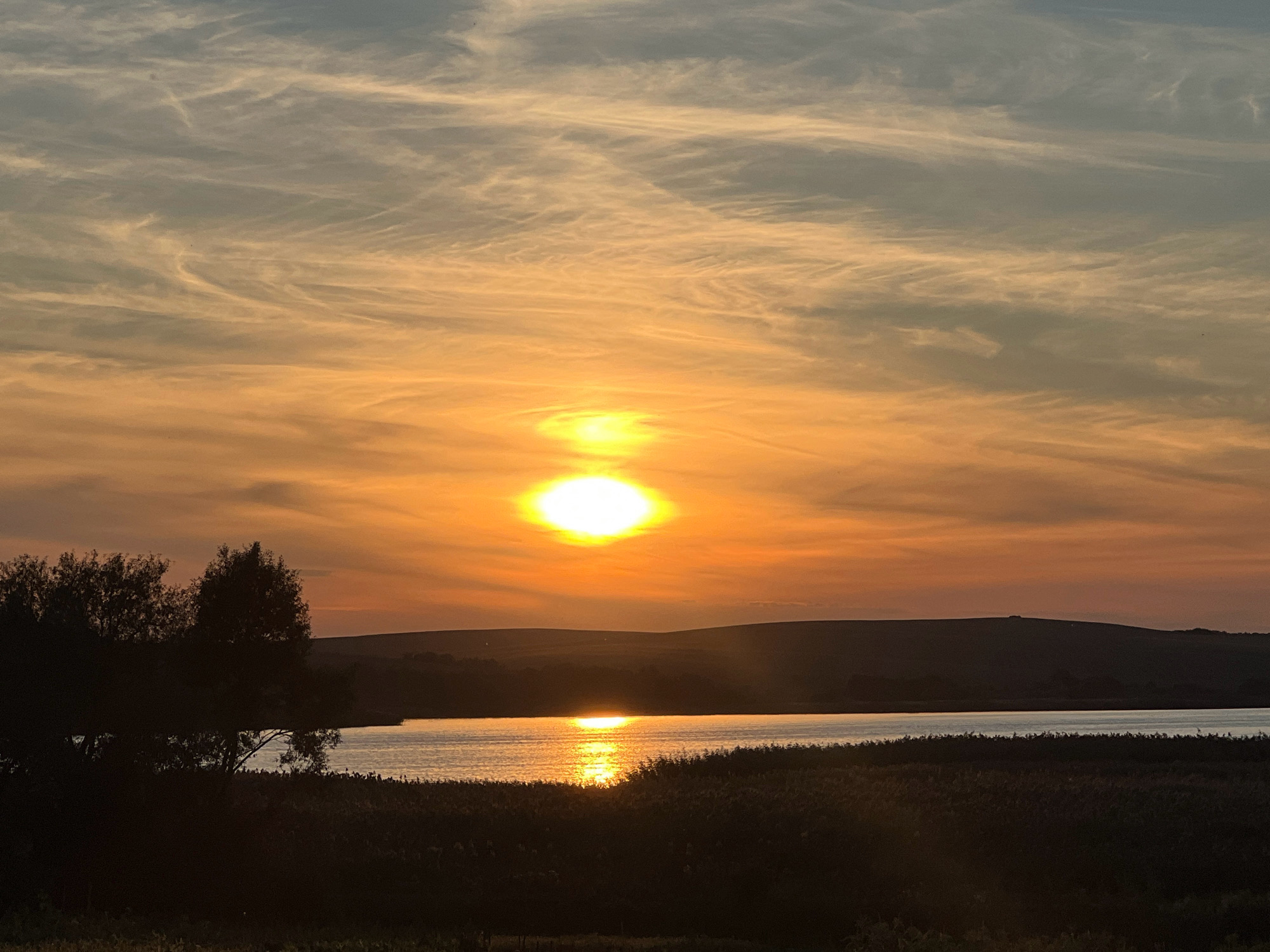 The image depicts a serene sunset over a body of water, likely a lake or river, surrounded by vegetation. The sky is awash with vibrant hues of orange, yellow, and blue, with wispy clouds streaking across. The sun, a brilliant orb of light, is positioned in the upper center of the frame, casting a shimmering reflection on the water's surface. Silhouetted against the warm glow of the sunset are rolling hills in the background and dark, dense foliage in the foreground. The overall scene conveys a sense of tranquility and natural beauty. The specific location in Vertelka, Ukraine, is not discernible from the image itself.