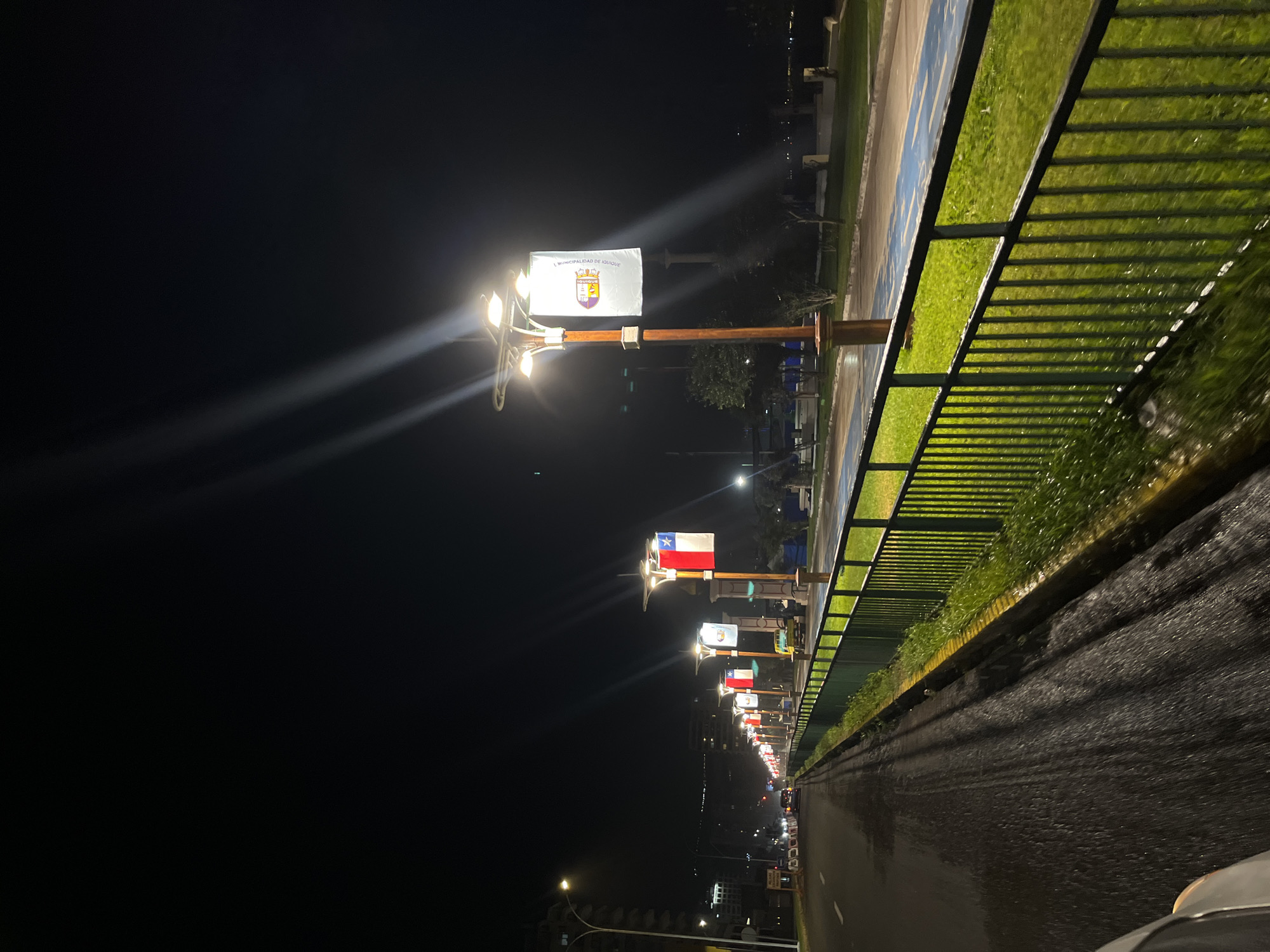 At night, a row of light posts lines a street in Iquique, Chile. Each post is topped with a light fixture and a flag, including the Chilean flag and the flag of the Municipality of Iquique. A metal railing separates the sidewalk and a grassy area from the road. The road is wet. Buildings can be seen in the distance.