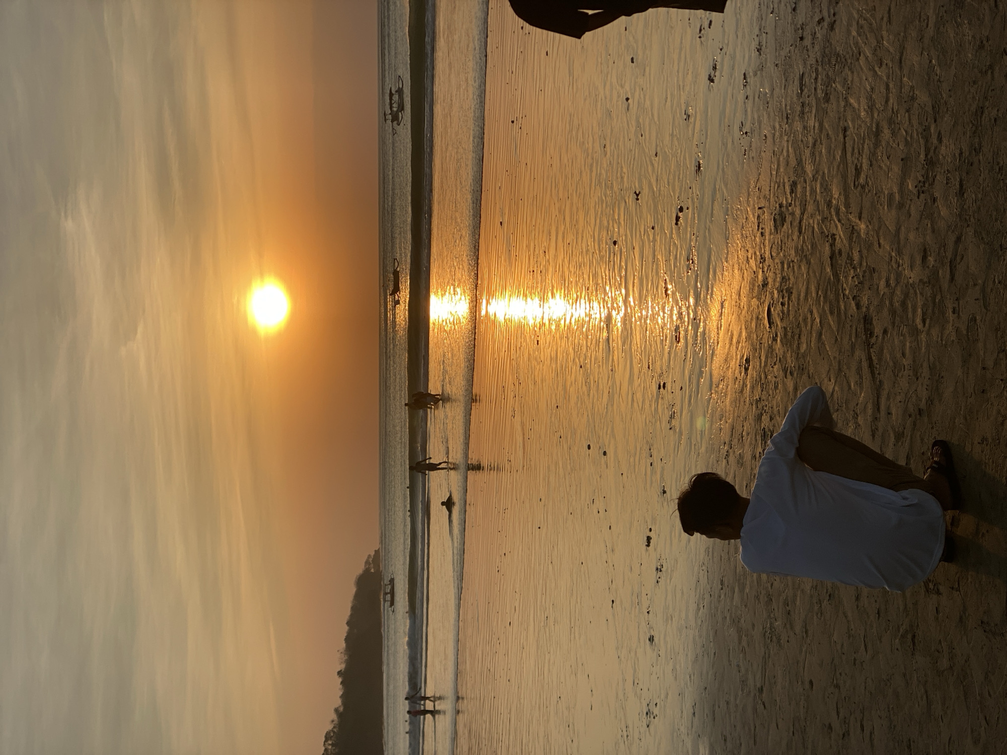 A wide-angle, vertical shot captures a sunset over the beach in Kuta Selatan, Indonesia. The sun, a bright orange orb, is positioned in the upper center of the frame, casting a shimmering reflection across the wet sand and gentle waves. In the foreground, a person kneels facing the sea, silhouetted against the warm glow. Further back, several other figures are visible wading in the water and walking along the shoreline. Small boats are scattered on the horizon, adding to the tranquil beach scene. The sky is a gradient of orange and yellow, with wispy clouds. The sand is textured with ripples and small dark pebbles.
