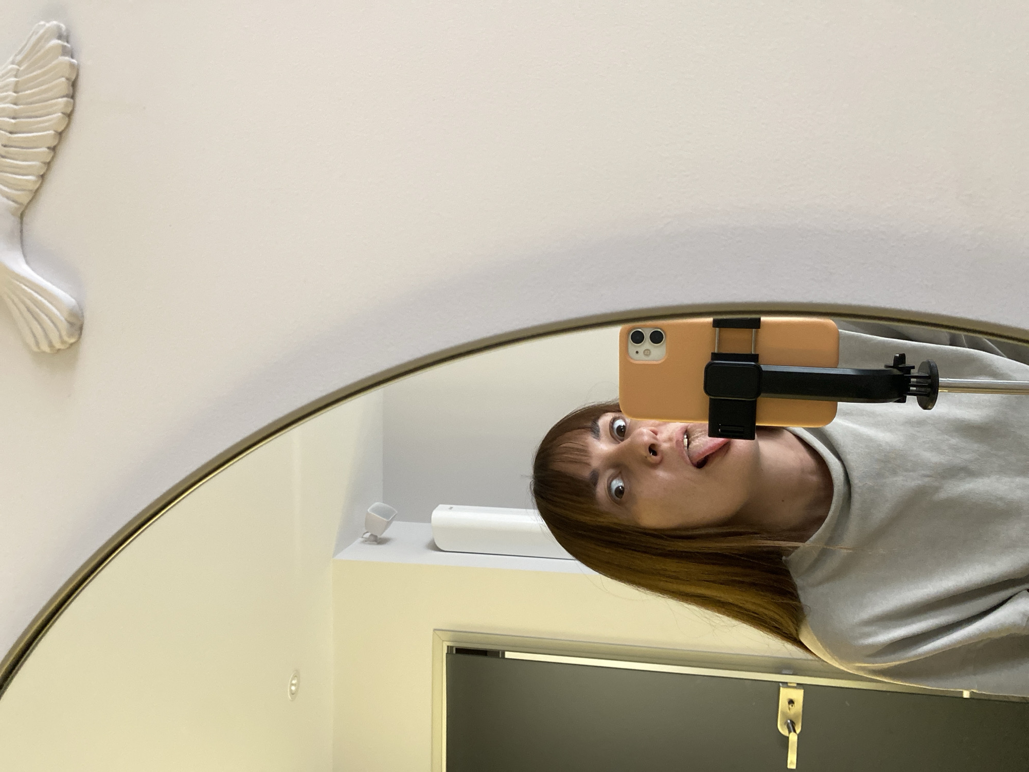 A selfie is reflected in a round mirror with a gold frame. The selfie shows a woman with brown hair and bangs sticking her tongue out. She is holding a phone attached to a selfie stick. The phone has a peach-colored case. In the background, a white wall with a decorative white wing is visible. Inside the mirror, a white shelf with a small object and a closed gray door with a gold handle can be seen. There is no information to determine if the location is in Votkinsk, Russia.