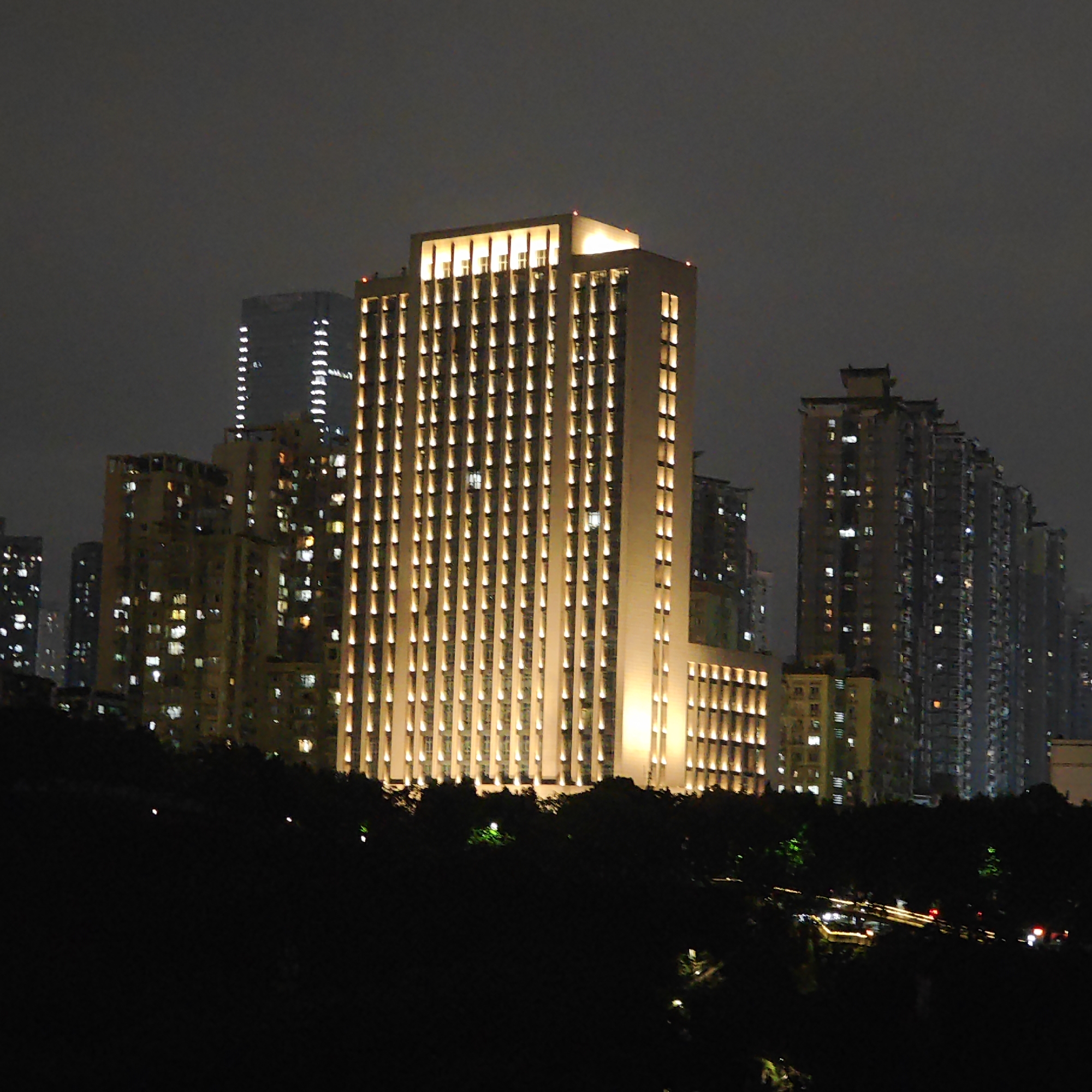 At night, a tall, illuminated building stands prominently in Yuzhong District, China. The building is rectangular, with vertical rows of lights accentuating its height. Other buildings of varying heights and illumination levels surround it. Below, a dark, silhouetted area likely indicates a park or wooded area with hints of paths and sporadic lights. A few distant vehicles can be seen, their headlights and taillights visible. The sky is dark and overcast.