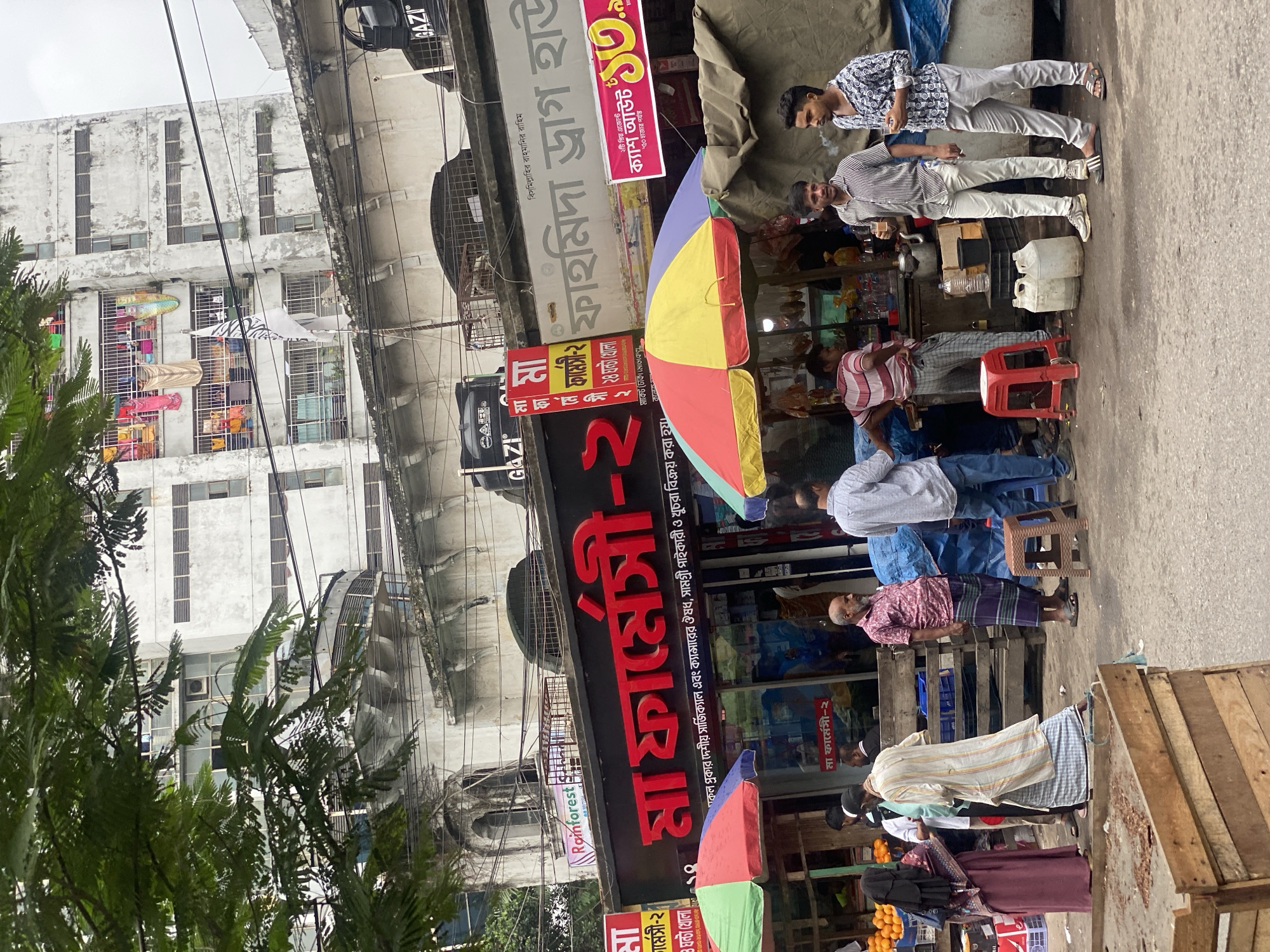 A street scene in Dhaka, Bangladesh, captures the bustling atmosphere of a commercial area. In the foreground, a wooden cart is partially visible, indicating a market or vendor setup. To the left, a woman in a maroon hijab and dress stands near a display of oranges. Several men are gathered, some in conversation, while others are engaged in activities related to small businesses. One man is seen with a cup of tea, and another is smoking.
Behind the street-level activity, a pharmacy with a prominent red sign reading "মা ফার্মেসী" (Ma Pharmacy) is visible. Adjacent to it is another sign that appears to be for a drug store. Colorful umbrellas provide shade over some of the vendors' stalls. In the background, a multi-story building with balconies and laundry hanging out to dry suggests residential occupancy above the shops. The architecture and the general appearance of the buildings are typical of urban areas in Dhaka. The sky is overcast, contributing to the muted light of the scene. The overall impression is one of everyday life and commerce taking place on a busy street in the city.