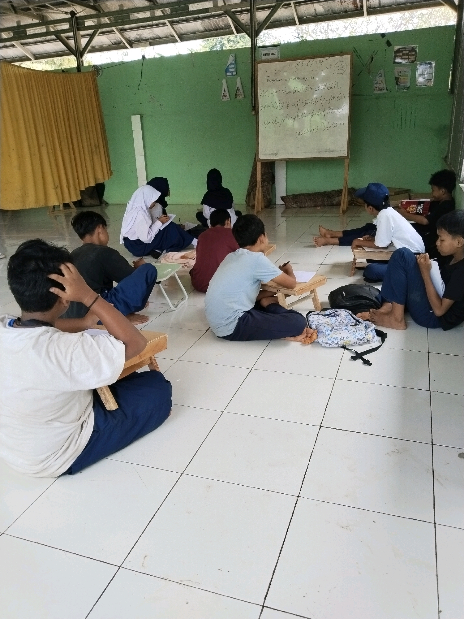 An overhead shot shows a group of students, mostly young boys and a few girls, sitting on the floor of a room in what appears to be a Madrasah Aliyah (Islamic Senior High School) in Serang, Indonesia. The room has a simple, open-air structure with a wooden beam ceiling and green walls. A whiteboard with Arabic writing is propped up on an easel. Some students are engaged in writing or reading, with papers and books scattered around them. Others are in a more relaxed posture, with one student scratching his head. The floor is tiled with a white and grey checkered pattern. In the background, a yellow curtain hangs on the left side of the room, and a fan is visible on the wall.