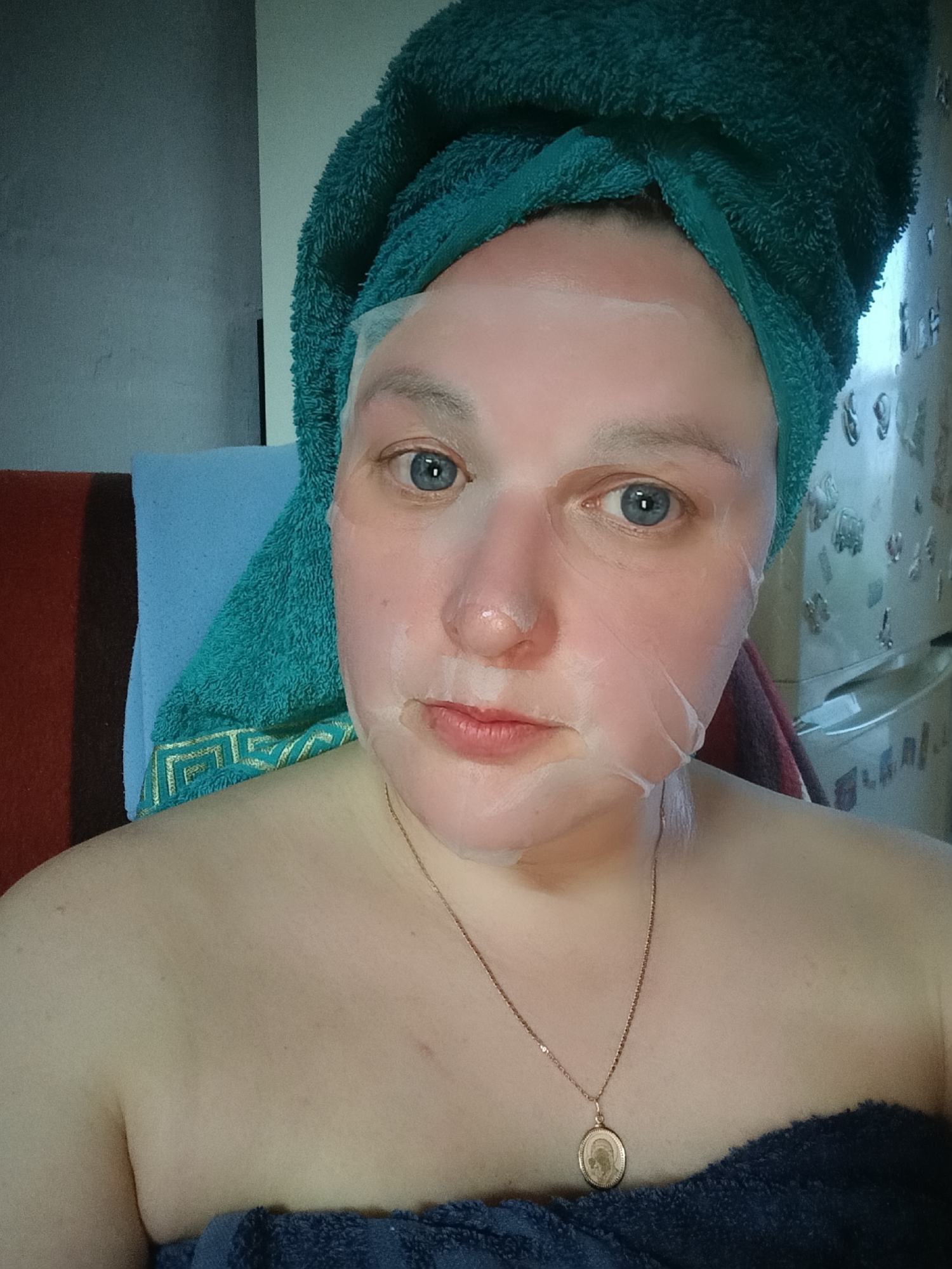 A woman is taking a selfie indoors. She is wearing a turquoise towel wrapped around her head and a sheet mask on her face. A gold necklace is visible around her neck. She is partially covered by a dark blue towel, leaving her shoulders bare. Behind her, there is a blue towel draped over a red-brown chair or blanket. A refrigerator with decorative magnets is visible in the background. The location is Chaikovsky, Russia.