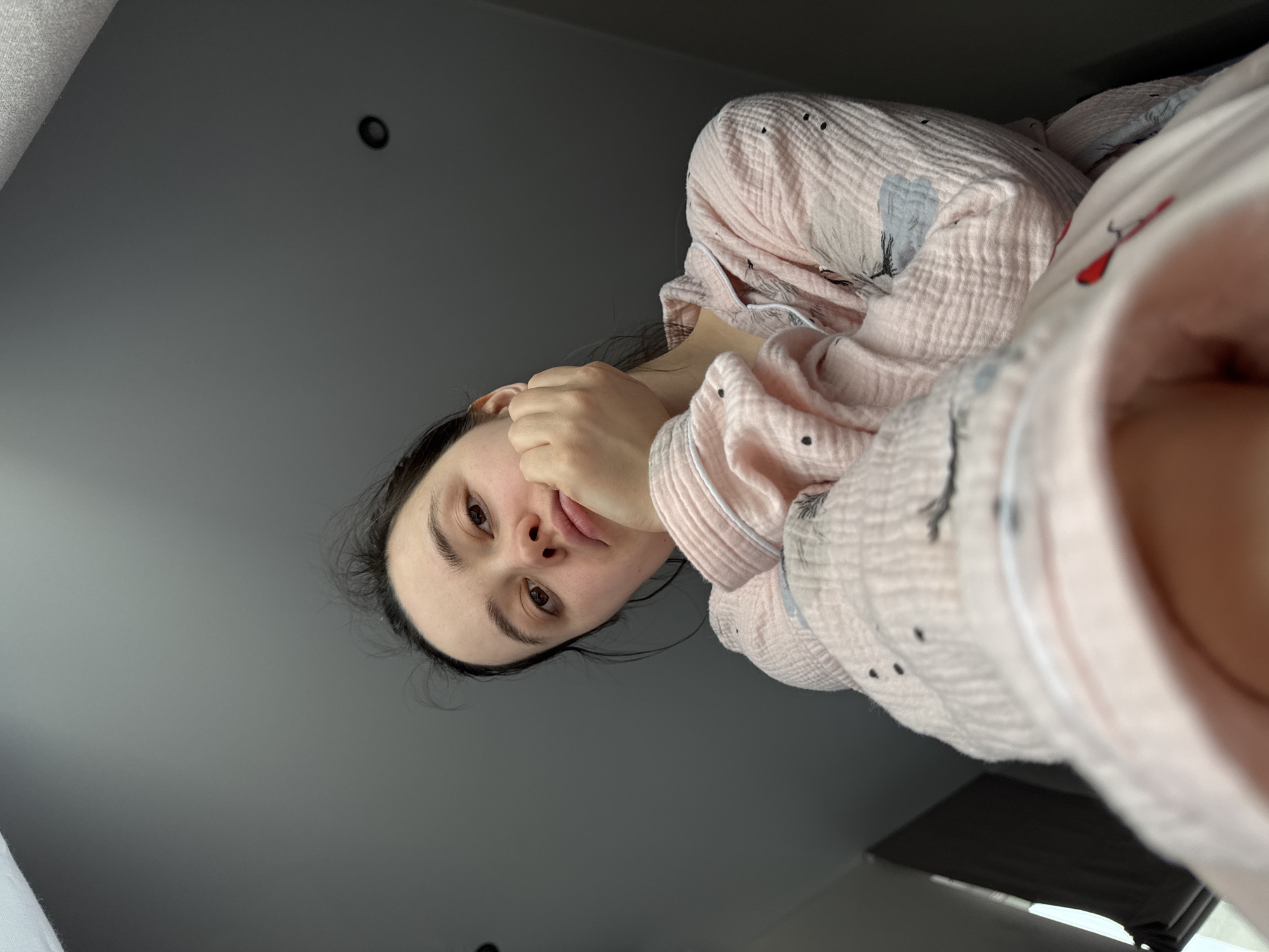 A woman with dark hair is looking down at the camera with a slightly pensive expression. She is wearing a pink and white patterned pajama top. Her hand is near her face, partially obscuring her mouth. The background is a gray ceiling with a small, round light fixture. The scene appears to be indoors. There are no elements identifying the location as Izhevsk, Russia.