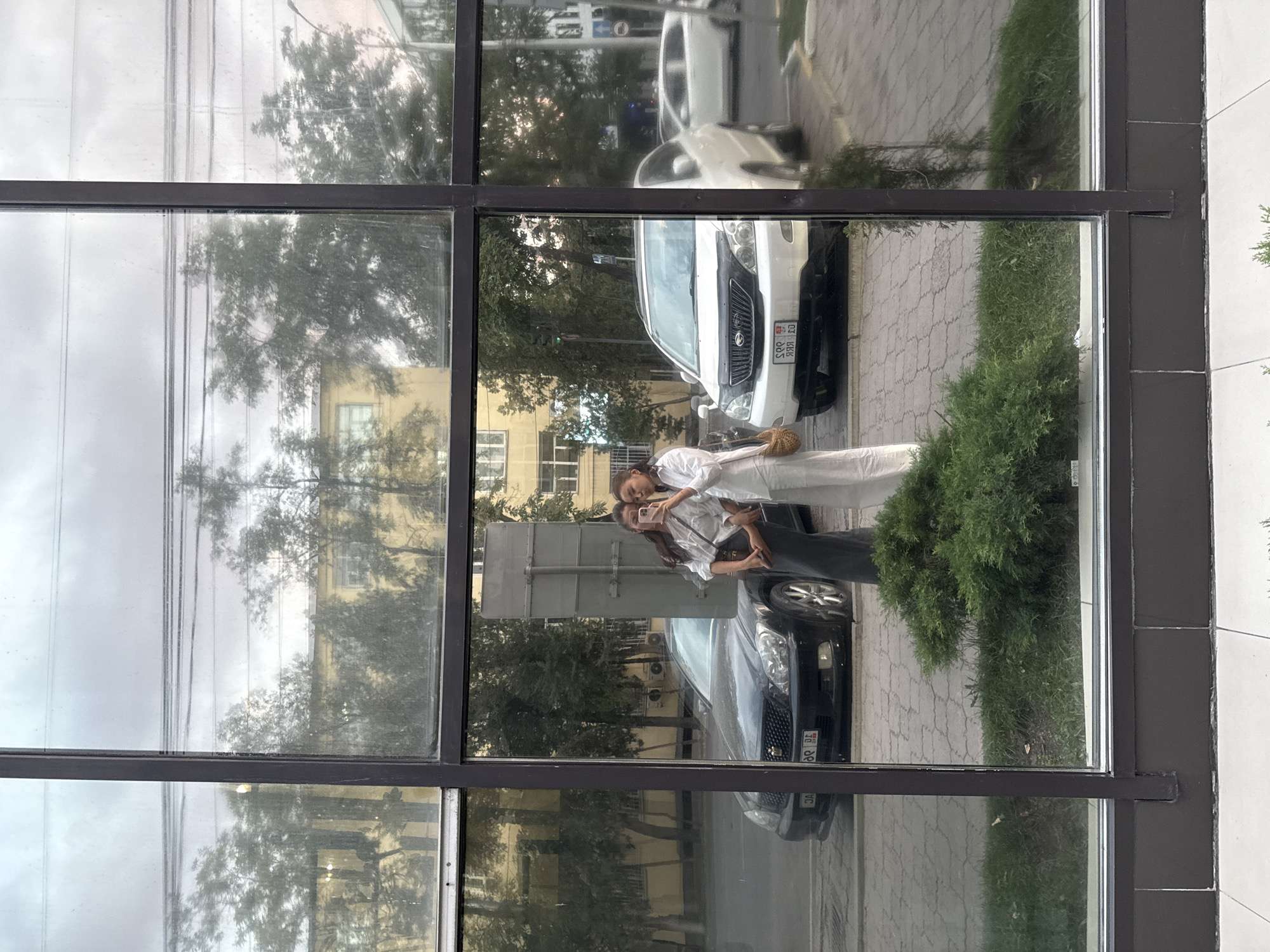 In Bishkek City, Kyrgyzstan, a photograph taken from a slightly low angle reflects two young women posing for a selfie in front of a large glass window. The women are dressed in casual, light-colored clothing. The woman on the left wears a white collared shirt and a dark, long skirt, carrying a tan crossbody bag. The woman on the right is in white pants and a white top, holding a smartphone up to take the picture. Their reflection is captured against a backdrop that includes a black sedan and a white SUV parked on a paved street. Beyond the vehicles, trees and a yellow building are visible, suggesting an urban setting. The foreground of the reflection shows a patch of green grass and a small, bushy evergreen shrub. The reflection is slightly distorted by the window's surface, which appears to be grimy. The sky above is overcast.