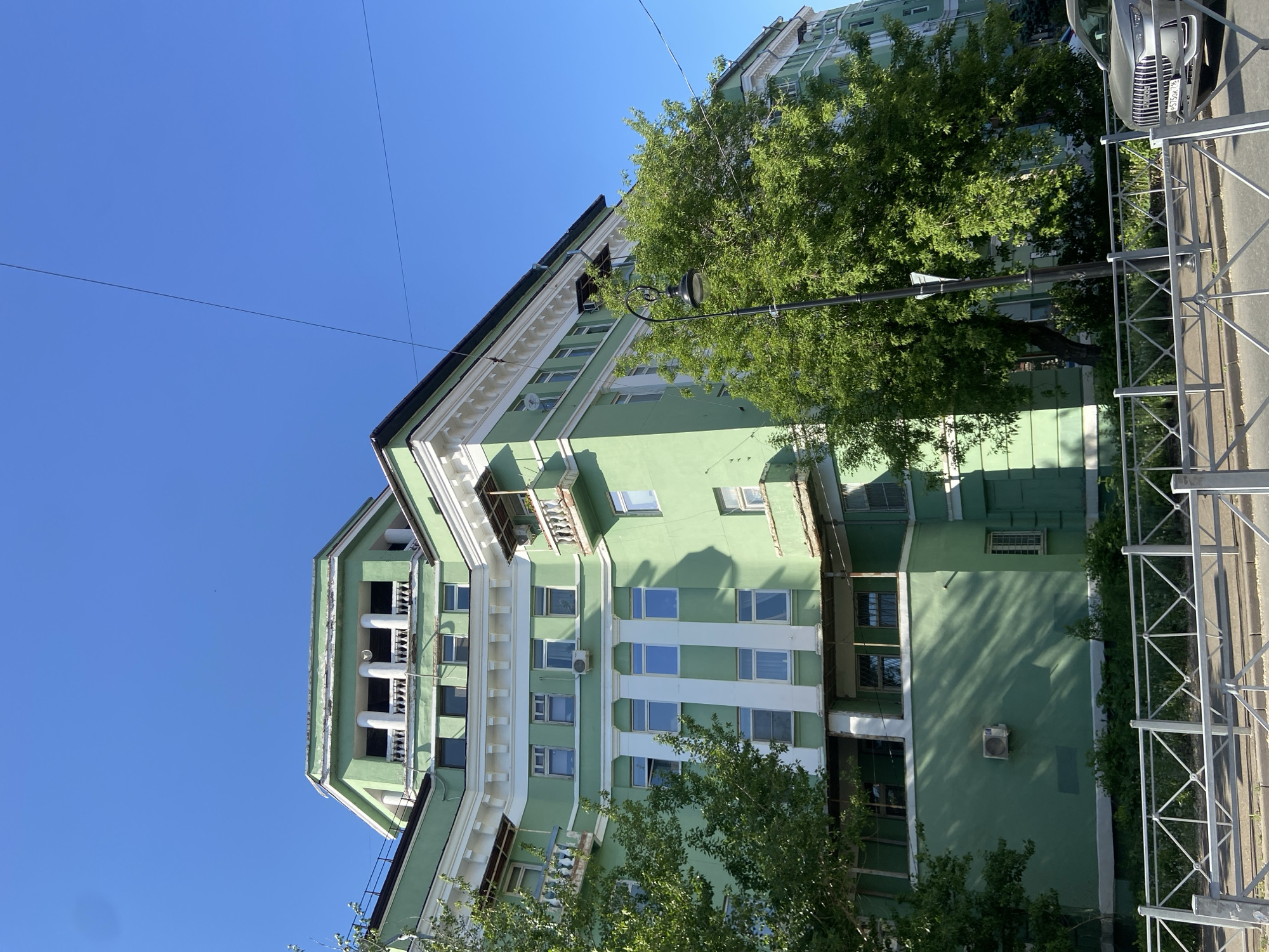The image shows a building in Kazan, Russia. The building is painted in green with white architectural details. It has multiple floors and windows, some of which are partially obscured by trees. A streetlight stands in front of the building, and a metal fence lines the sidewalk. A car is parked on the right side of the frame. The sky is clear and blue.