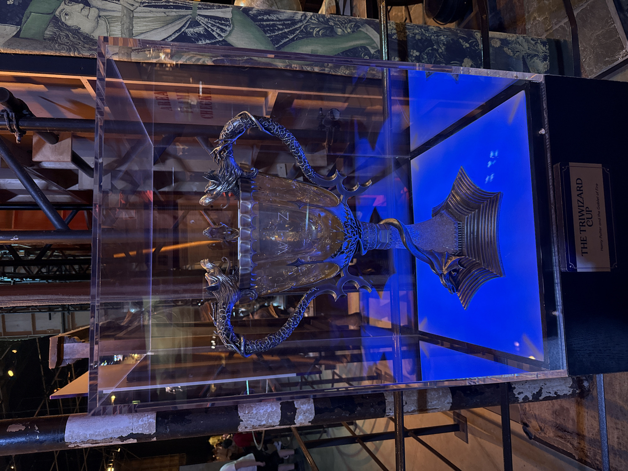 The image displays a display case containing the Triwizard Cup from Harry Potter and the Goblet of Fire. The cup is made of metallic material and features dragon-shaped handles. It is illuminated from below by a vibrant blue light, which reflects off the transparent casing. The display case is situated on a dark, rectangular base with a plaque on the front that reads "THE TRIWIZARD CUP Harry Potter and the Goblet of Fire".
The background of the image is somewhat blurred, but it appears to be an indoor exhibition or museum setting. Visible in the background are industrial-looking structures, metal scaffolding, and a tall, dark pillar on the left side of the frame. There are also glimpses of people in the distance, suggesting a public space. To the right of the display case, a large tapestry or banner with a medieval-style illustration of a person in a green hat is visible. The floor appears to be a stone or tile material.
The context of "Three Rivers, United Kingdom" is not visually evident in the image itself.