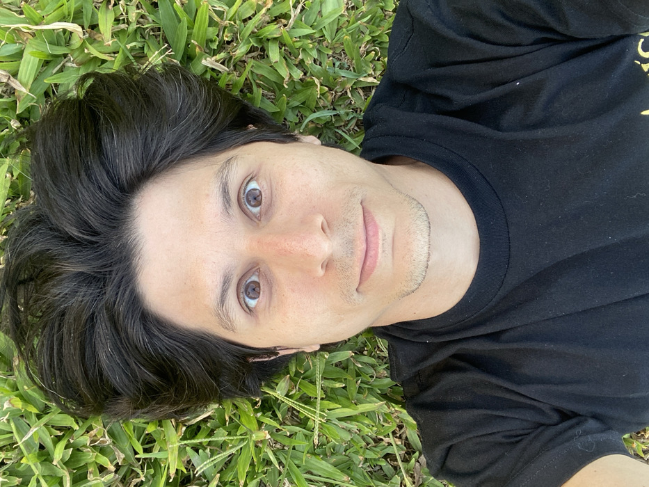 A man is lying on his back on green grass, captured from a top-down perspective. He looks directly up at the camera with wide, brown eyes and a subtle, relaxed smile. His dark, medium-length hair is fanned out around his head on the grass. He has fair skin and a faint stubble around his chin and mouth. He is wearing a plain black t-shirt. The background is entirely composed of vibrant green grass, suggesting an outdoor setting like a park or garden. The lighting is natural and bright, indicating it is daytime. The overall mood conveyed by his expression is calm and serene. The location is implied to be outdoors in Buenos Aires, Argentina. No text is visible in the image.