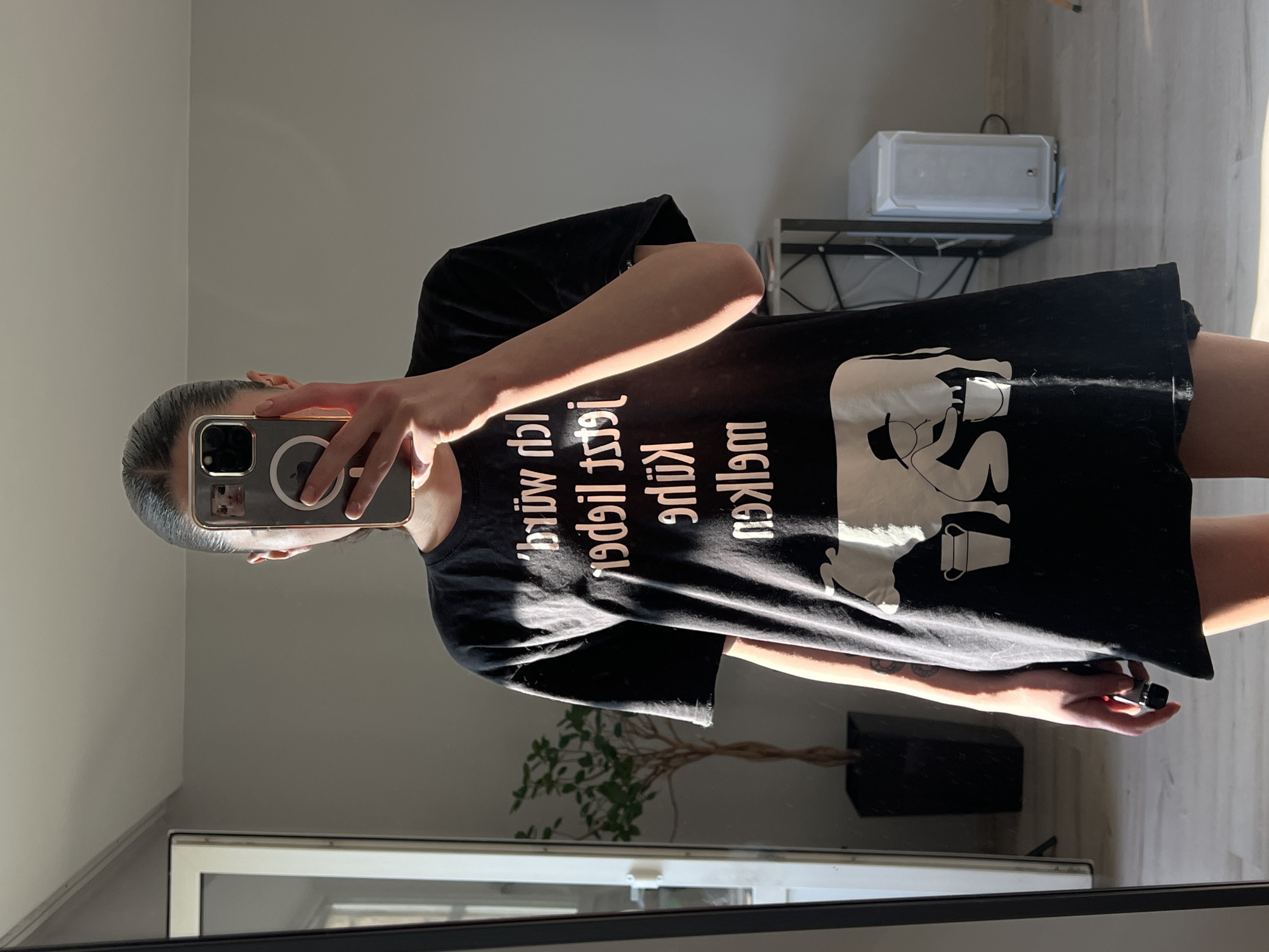 A person stands in front of a mirror, taking a selfie. They are wearing a black t-shirt with white text and a graphic of a person milking a cow. The text on the shirt appears to be in German and reads "Ich würd' ja gern aber jetzt lieger Kühe melken" which translates to "I'd love to but I'd rather milk cows." The person is holding a smartphone in their right hand, positioned to capture their reflection. Sunlight streams in from the left side of the frame, creating strong highlights and shadows on the person and the surrounding environment. The background includes a mirror, a doorway, a plant in a pot, and furniture including a computer tower. The scene appears to be an indoor residential setting. The location is identified as Ternopil, Ukraine.