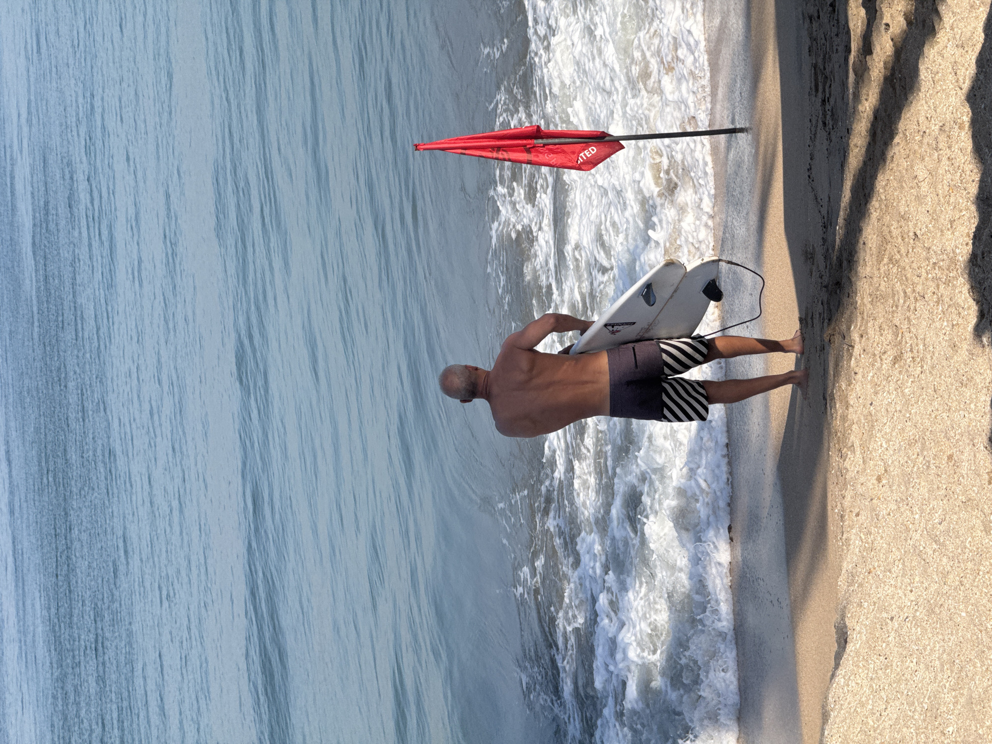 A man stands on a sandy beach in Kuta, Indonesia, holding a white surfboard. He is facing the ocean, which is calm with gentle waves. A red flag is planted in the sand to his right, indicating dangerous conditions or a restricted area. The man is wearing dark swim shorts with white stripes. The beach is mostly empty, with the exception of the man and the flag. The water is a clear blue, and the sand is light brown. The sky is clear and the sun is shining.