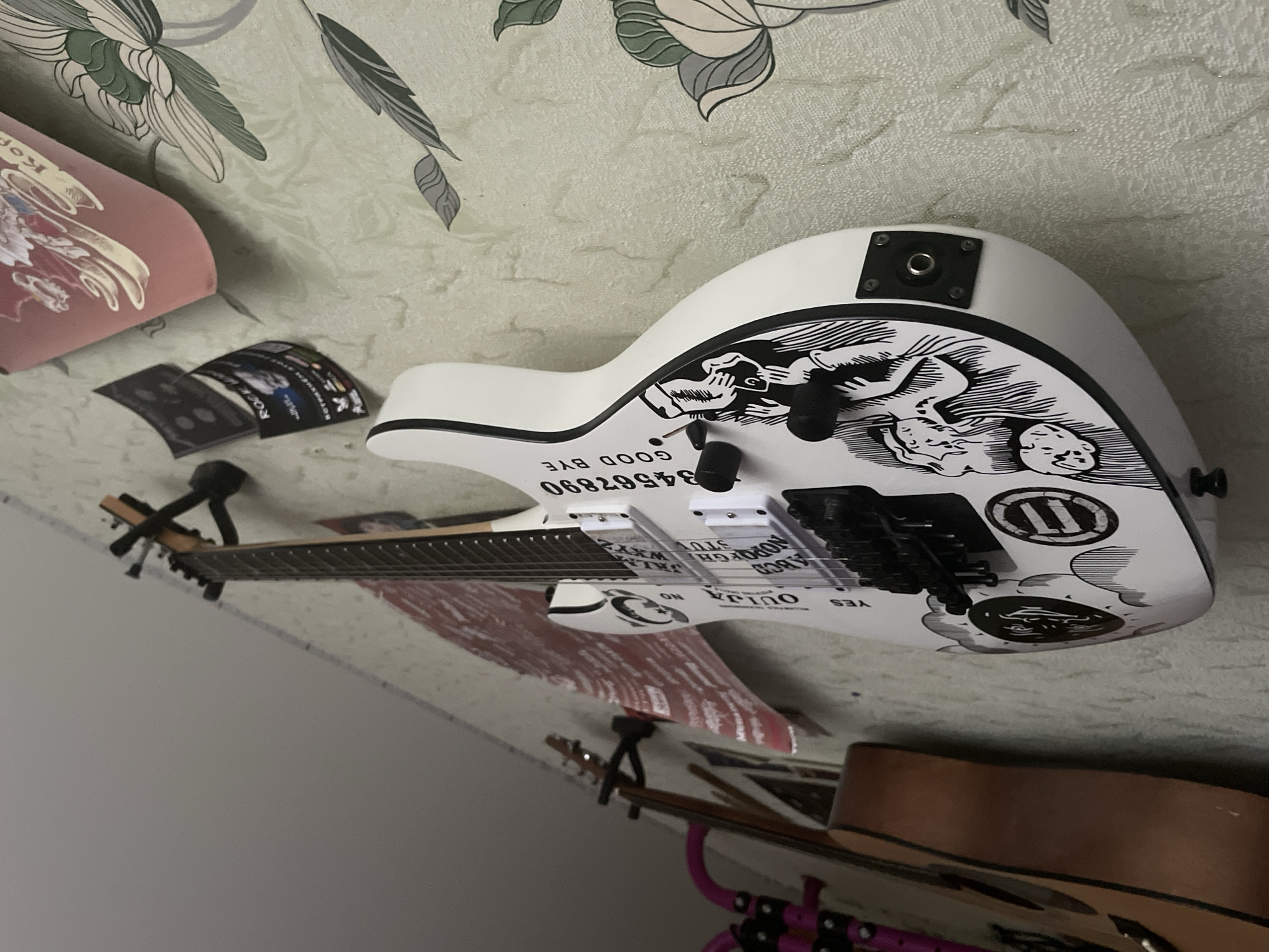 This image captures an indoor scene, likely a room located in Chaikovsky, Russia. The background features light-colored, textured wallpaper adorned with a decorative pattern of green and grey leaves.
The central subject is a white electric guitar, distinguished by intricate black and white graphics that mimic a Ouija board design. The guitar's body showcases a large, stylized illustration of a creature or demon. Visible text and symbols on the guitar include "GOOD BYE", "34567890", "OUJIA", "YES", "NO", and segments of the alphabet "ABCDEFG WXYZ". The guitar is fitted with black hardware, including pickups, bridge, control knobs, and an output jack.
Other objects in the scene include a pink item in the upper left, possibly a pillow or cushion, bearing a partial graphic with the Cyrillic text "КОРО". Below this, two small, dark rectangular cards are visible; one clearly displays "Rock N Roll" and some additional Cyrillic text. A small white object, possibly a computer mouse, is partially visible near these cards. In the lower part of the frame, a portion of a brown acoustic guitar is seen, alongside what appears to be another guitar or stand with a pink/purple element.
No specific activity or event is taking place; the objects are stationary, suggesting they are part of a personal collection or display within the room. The image itself is rotated 90 degrees clockwise.