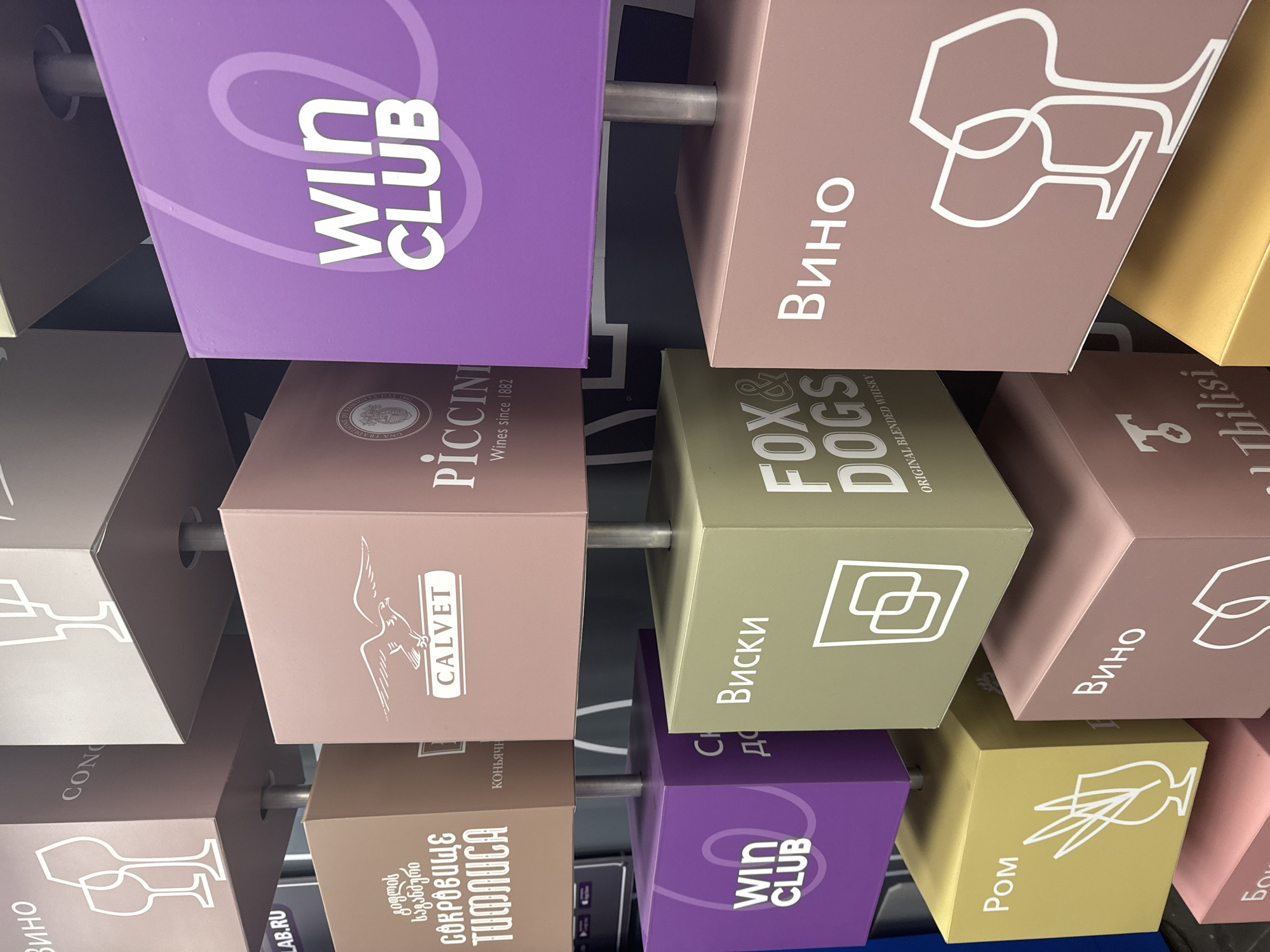 The image shows a display of rectangular boxes suspended from a central metal rod, possibly in a store or exhibition setting. Each box has different branding or labels related to alcoholic beverages.
The boxes show the following brands and labels: "Win Club", "Вино" (Russian for Wine), "PICCINI Wines since 1882", "CALVET", "СОКРОВИЩЕ ТИФЛИСА" (Treasure of Tbilisi, in Russian), "FOX & DOGS Original Blended Whisky", "Виски" (Whisky in Russian), "Ром" (Rum in Russian), "Tbilisi". There are additional markings with Cyrillic text and graphics that indicate the type of drink.
The scene likely takes place in Moscow, Russia due to the presence of Cyrillic text and the promotion of drinks associated with Russian culture and language.