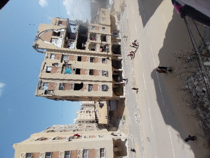 An elevated view captures a street scene in Khan Yunis, Palestinian Territory, under a clear blue sky with scattered clouds. The foreground shows a debris-strewn edge, with the shadow of an unseen structure extending across a partially marked paved road. Several individuals are present: a group of approximately five adults and children walk on the right side of the road, one pushing a stroller; two more individuals walk further left on the road, casting long shadows. On the right, a multi-story residential building, light tan in color, exhibits severe structural damage, with extensive portions of its upper floors collapsed or missing, revealing internal spaces. A distinctive arch-shaped architectural feature on its upper right is partially destroyed. Numerous windows on this building are shattered or absent. A blue tarp hangs from one window opening. To the left, another similarly styled multi-story building shows less severe but still visible damage, including broken windows, and a red fabric item hangs from a balcony on its left side. Further back, additional damaged residential structures are visible. Rubble and debris are scattered on the ground surrounding the buildings and along the road shoulders. Several individuals are also seen walking near the bases of the buildings.