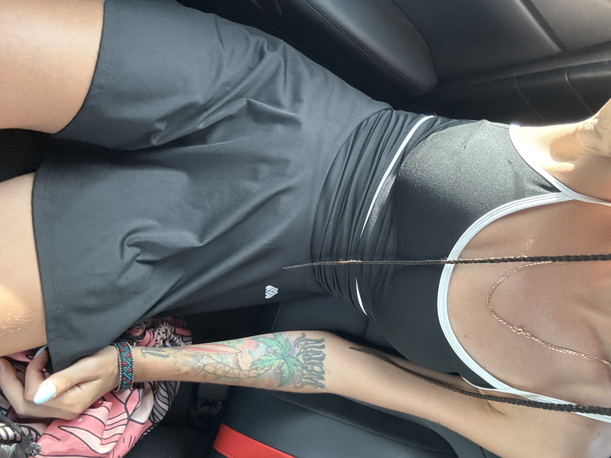 The image shows a close-up of a person's torso and arm, sitting in a car. The person is wearing a black athletic top with white trim and a black skirt. They have braided hair that extends down their chest, and a gold chain necklace. The person's left arm is visible, featuring a colorful tattoo of a palm tree and other imagery, as well as a beaded bracelet. The background is dark, consistent with the interior of a car, with a hint of a red seatbelt. The scene is not identifiable as being in Hoa Lư Ward, Vietnam.