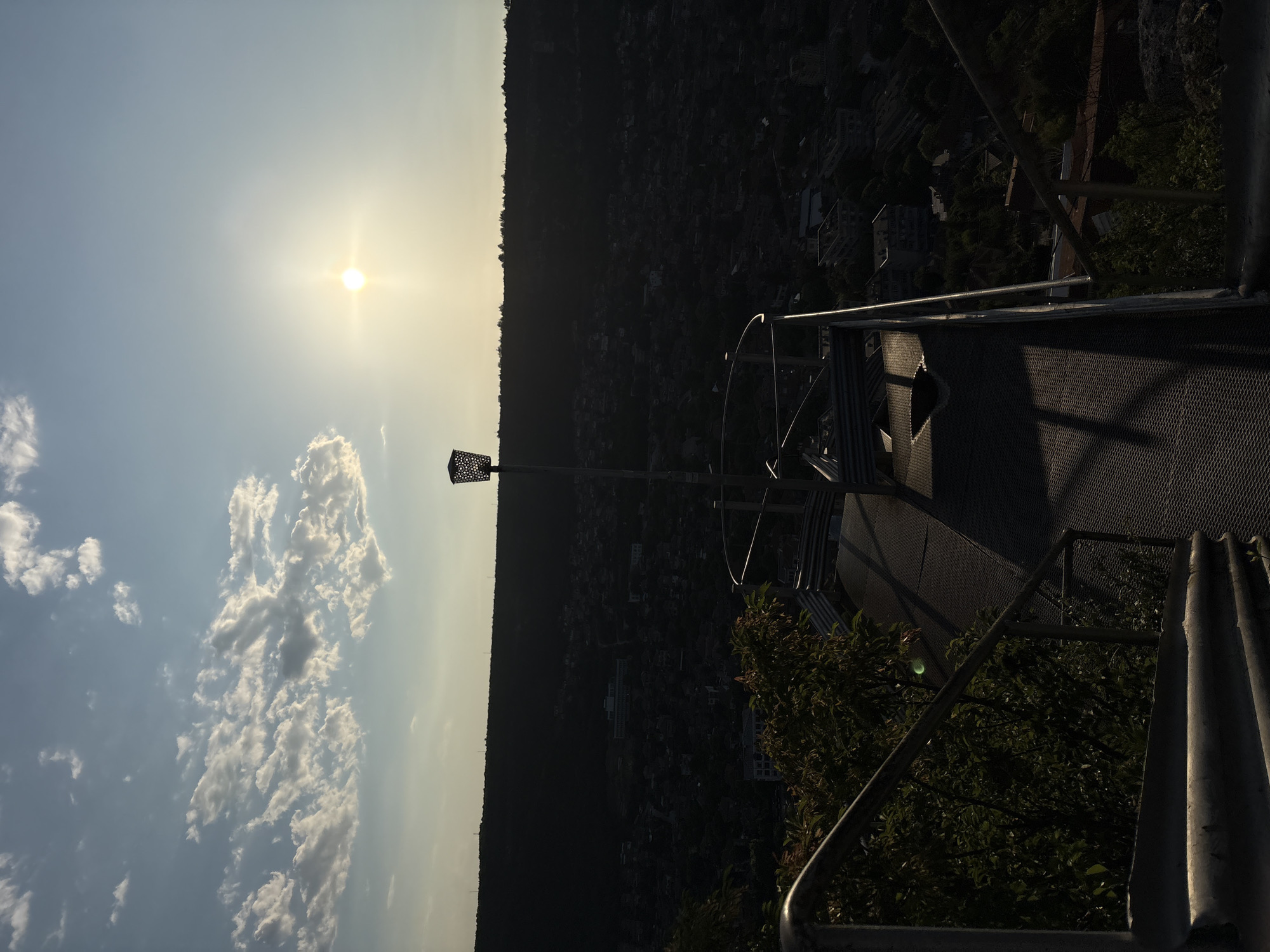 The image depicts a panoramic view of the city of Provadia, Bulgaria, observed from an elevated metallic viewing platform at either sunset or sunrise. The prominent, bright sun with a starburst effect is positioned in the upper right quadrant of the sky, casting long shadows and creating a strong backlighting effect on the landscape. The sky features scattered white cumulus clouds, primarily on the left side.
In the foreground, a metallic platform with corrugated and grated sections is visible, equipped with silver railings. A distinct tall, thin lamp post, topped with a decorative, perforated lantern-like fixture, stands on the platform, slightly right of center. Dark green foliage from bushes or trees is visible on the left side of the platform, partially obscuring the view immediately below.
The midground reveals the city of Provadia, sprawling across the valley below. Due to the intense backlighting from the sun, the urban area appears largely in silhouette or deep shadow, showing numerous residential and commercial buildings. A larger, lighter-colored building stands out amidst the darker structures in the central-left midground.
The background consists of a dark, undulating treeline of a forested hill or mountain range, forming the horizon line above the city. No individuals or discernible actions are present within the frame.