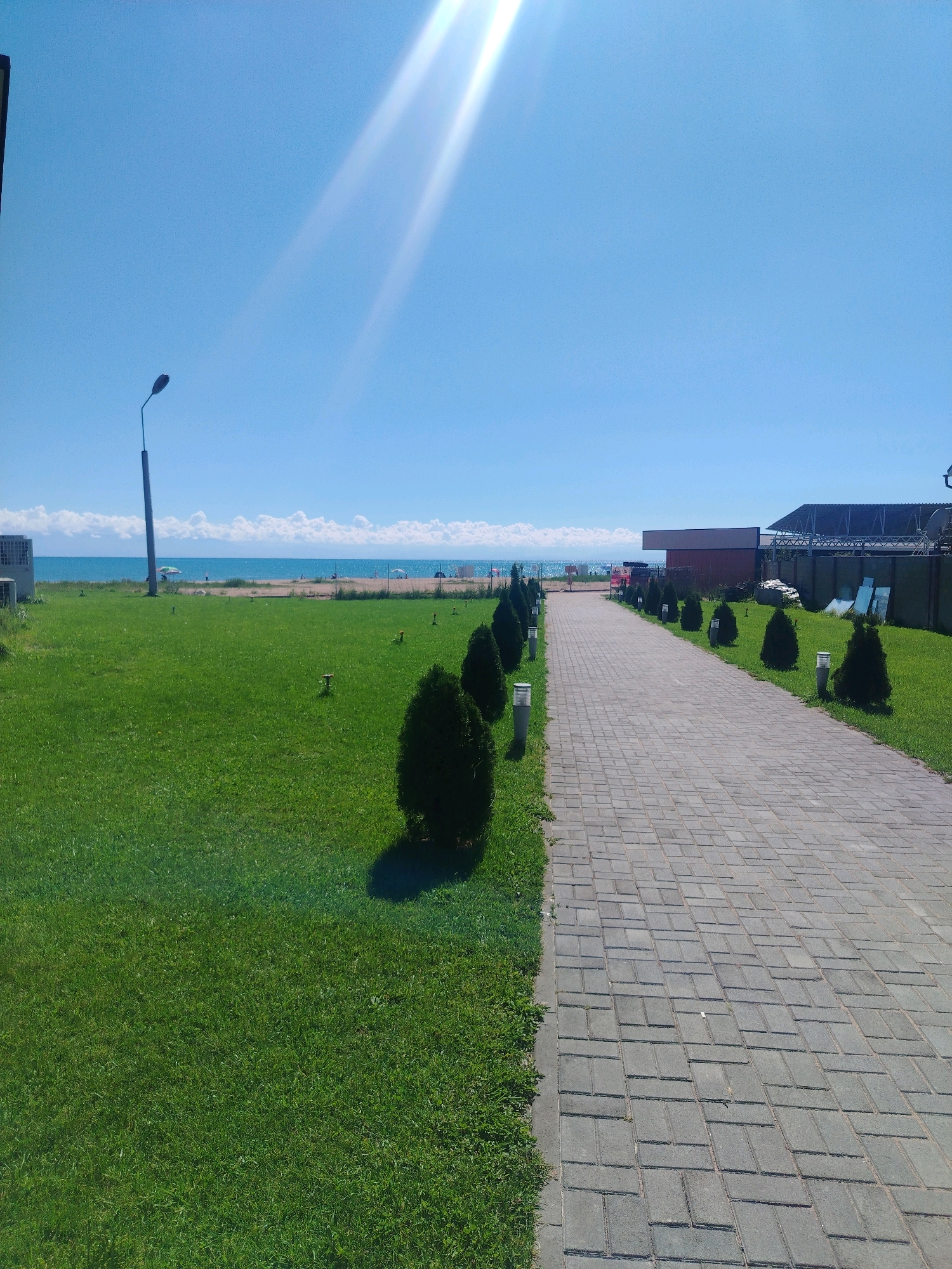 A paved pathway with grey rectangular stones leads away from the viewer towards a body of water, likely a lake, under a clear blue sky. The pathway is bordered on the left by a well-maintained green lawn with several small, cone-shaped evergreen trees and low-lying lights. On the right side of the pathway, there is also a green lawn, more evergreen trees, and a dark fence topped with a covering, behind which are structures. In the distance, beyond a strip of sandy beach, is the tranquil blue water. The sun is bright and casts strong light and shadows, creating a glare in the upper right quadrant of the sky. The scene appears to be located in Bosteri, Kyrgyzstan, possibly a resort or recreational area given the proximity to the water and the organized landscaping.