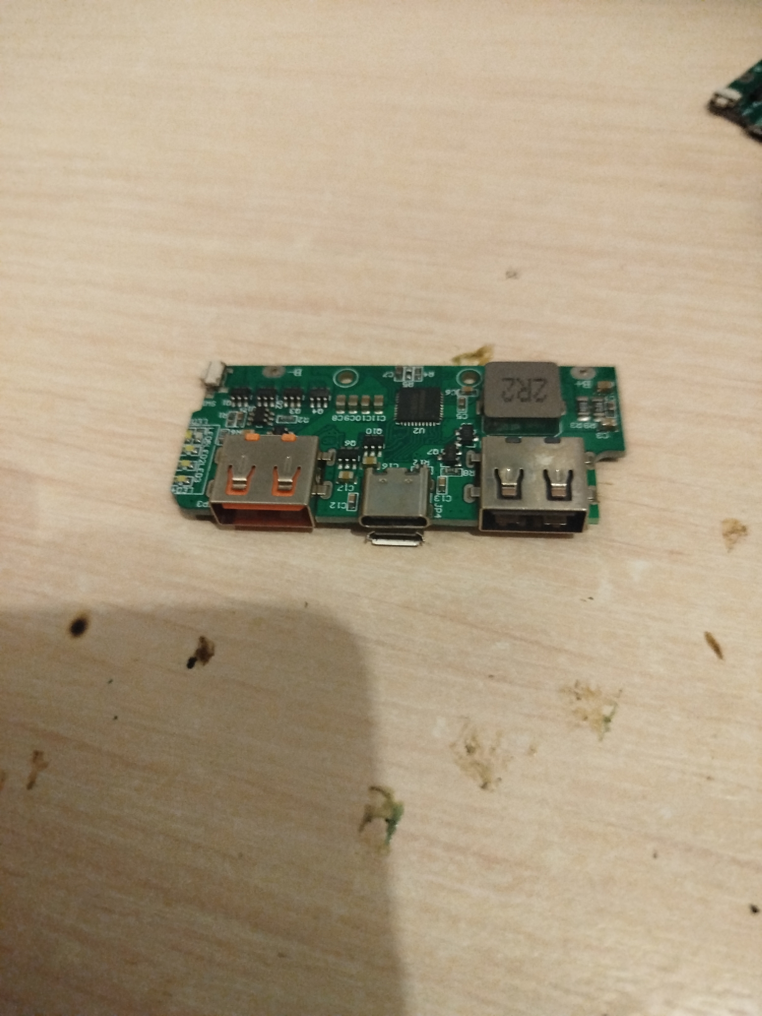 The image shows a close-up, overhead view of a green circuit board with various electronic components. The circuit board is placed on a light-colored, wooden surface. Visible components include several USB ports (one orange USB-A and two silver USB-A ports), a USB-C port, and a black rectangular component labeled "2R2". There are also smaller black integrated circuits, resistors, and capacitors. The lighting creates shadows on the wooden surface. The scene does not depict any people, actions, or interactions. No geographical markers of Verkhnodniprovsk, Ukraine, are present in the image.