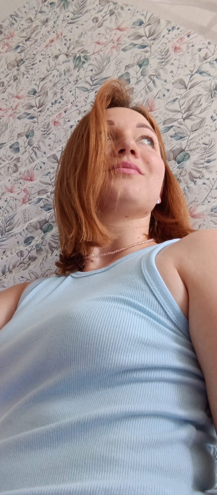 The photograph presents a low-angle view of a fair-skinned woman with auburn hair. Her head is tilted upwards and slightly to the right. She is wearing a light blue ribbed tank top and a delicate pink beaded necklace. A small stud earring is visible in her right ear. The background consists of a floral wallpaper with a muted color palette of blues, greens, grays, and pinks. The location is not discernible, and therefore cannot be placed in any city.