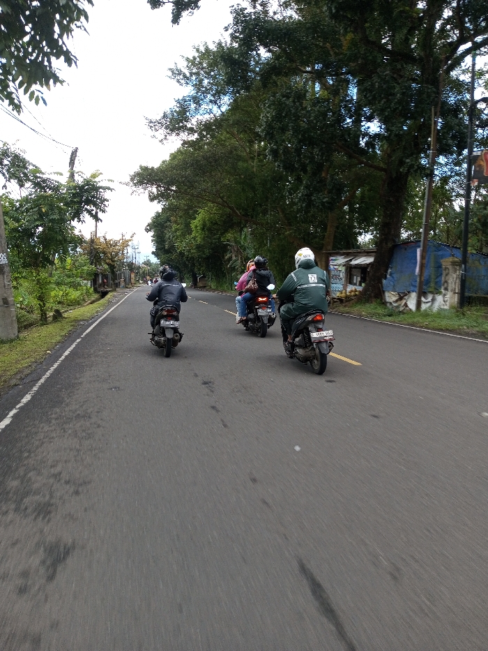 The image depicts a view along an asphalt road in Serpang Shalli, Bhutan, under an overcast sky. Three individuals are riding motorcycles away from the viewer. From left to right: a solo rider in a dark jacket and dark helmet on a dark scooter; a second scooter with two occupants, the rear passenger wearing a pink top and dark helmet; and a solo rider in a green jacket with a white logo on the back, wearing a white helmet, on a dark scooter with license plate "F 5885 VLC". The road surface shows dark stains and is bordered by a white line on the left. The left side of the road features a grassy verge, a concrete utility pole with "380/200" and "02" visible, and dense green foliage. The right side of the road is lined with large trees, under which a makeshift structure of corrugated metal and blue tarpaulin is situated, possibly a stall, with light-colored objects visible in front. A concrete pillar stands next to this structure.