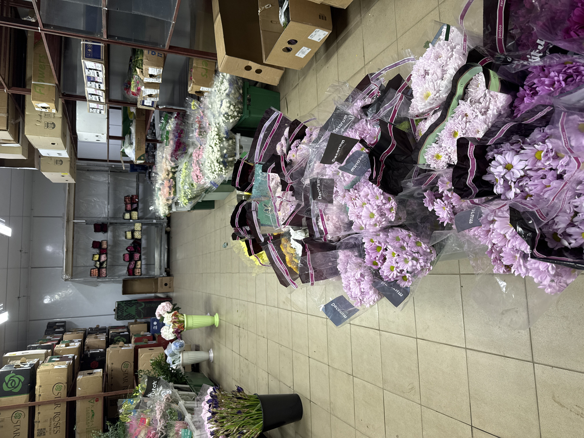 The image captures the interior of what appears to be a flower storage or wholesale facility, possibly located in Izhevsk, Russia given that the OCR detected "Nothing like it!," which is a rose brand registered in Russia.
The scene is dominated by rows of flowers, mostly in individual plastic wraps and arranged in groups. Prominent among them are bouquets of purple and white daisies, easily identifiable by their round shape and contrasting centers. The wrapping around each bouquet is secured with black and purple bands labeled "zentoo" and "Decorum."
Further back in the space, there are more flowers in various colors and stages, some arranged in vases. The setting includes shelves laden with cardboard boxes, some labeled "Flowers" and "STAR ROSES." The floor is tiled in a neutral color. The facility appears organized with an industrial shelving system along the walls. No people are visible.