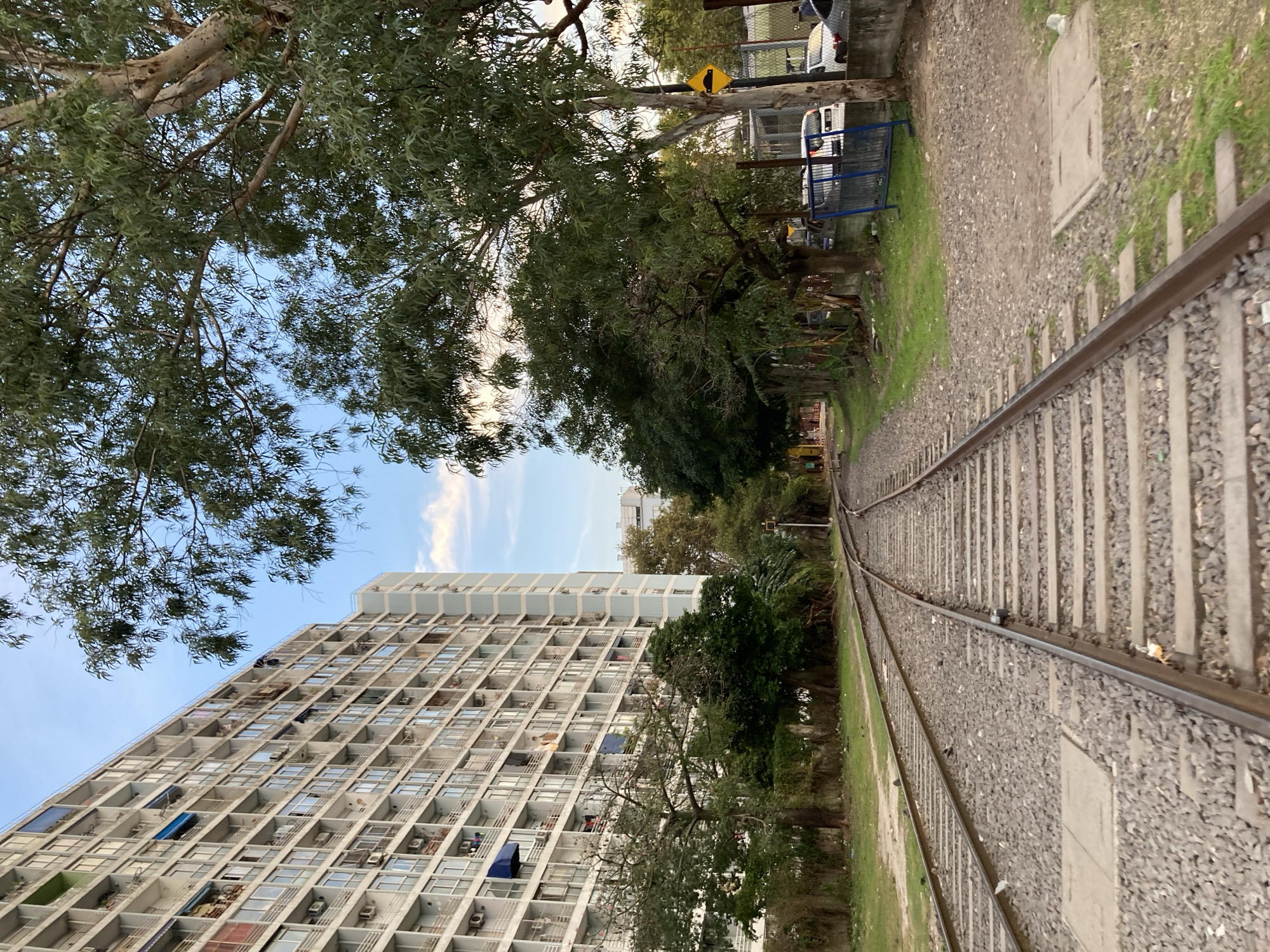 A low-angle shot presents a railway track stretching into the distance, flanked by trees and greenery. A tall, rectangular building, densely populated with windows and air conditioning units, stands on the left. The sky is visible through the branches of the trees, displaying a blend of blue and white clouds. The railway tracks are lined with gravel and weeds, and a street sign can be seen among the trees. The photograph is taken in Buenos Aires, Argentina, where these types of buildings can be found.