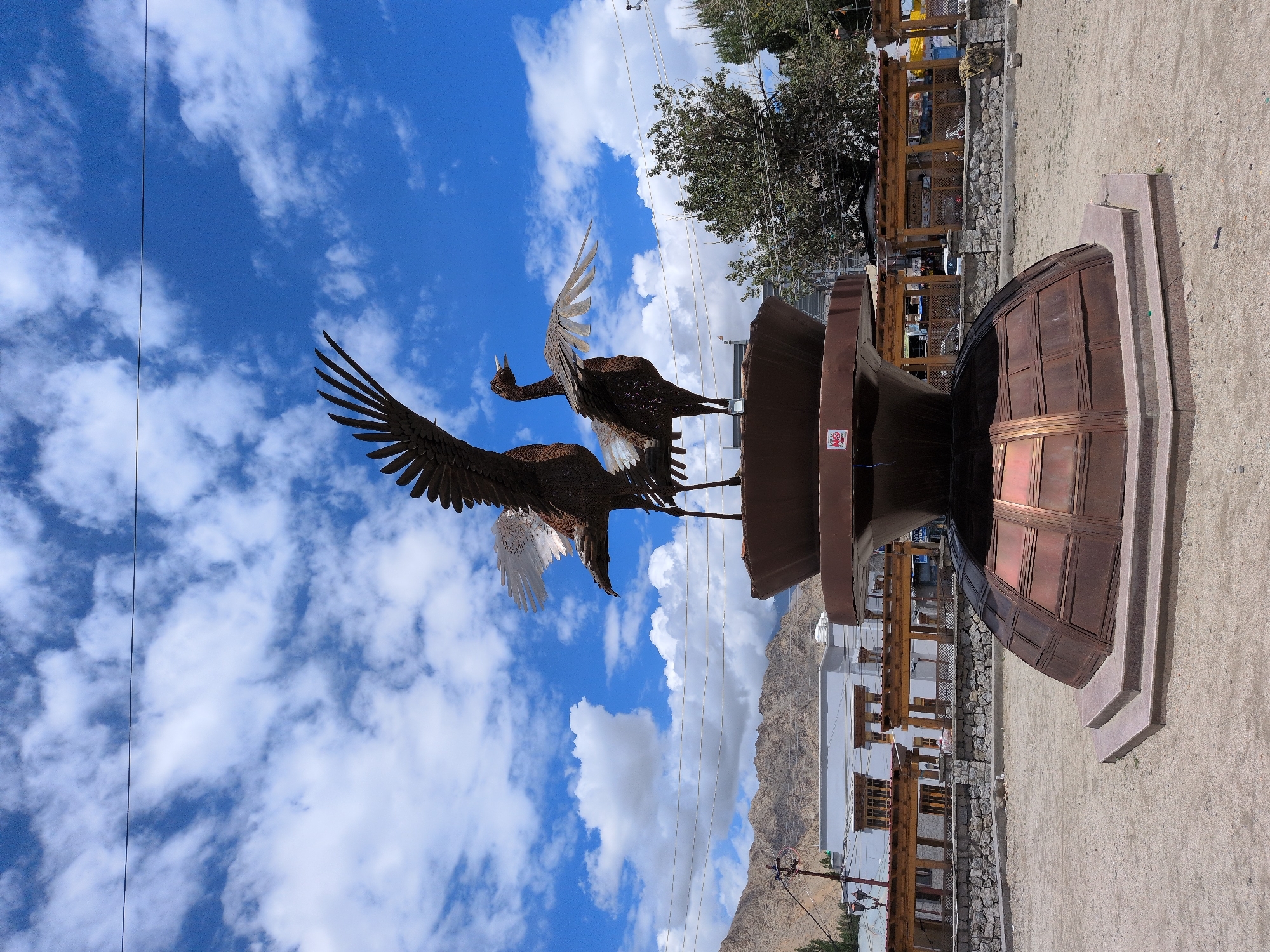 The image shows a close-up view of a large metallic sculpture of two cranes with their wings spread, positioned on top of a tiered, bell-shaped pedestal. The sculpture is situated in an open, dusty area, likely a public square or park in Leh, India, given the arid, mountainous landscape in the background and the traditional architecture of the buildings visible behind the sculpture. The sky is a vibrant blue with scattered white clouds, and power lines are strung across the upper part of the frame. The sculpture itself appears to be made of dark metal, with some lighter, reflective accents on the wings. The pedestal is a darker, reddish-brown metallic color with segmented panels. In the distance, there are low-rise buildings with wooden accents and covered walkways, possibly shops or market stalls, and a few trees. The ground is a mix of dirt and gravel, with some scattered debris. The overall scene suggests an outdoor public space in a somewhat arid, high-altitude region.