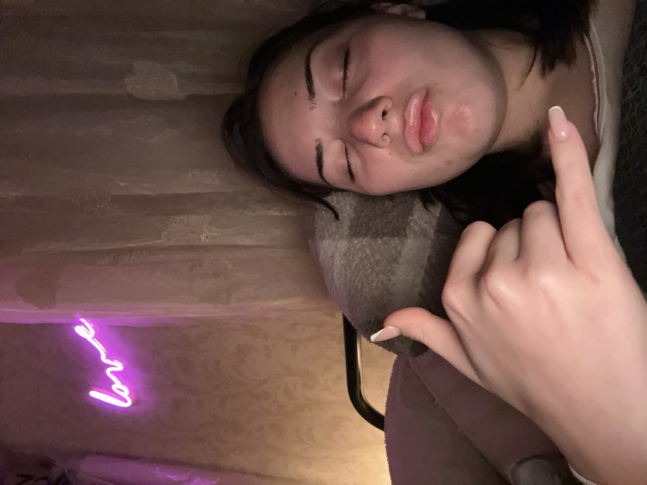 A woman lies with closed eyes, resting her head on a gray plaid pillow. A hand is raised, fingers extended with the middle finger prominently displayed. The background consists of a patterned surface with a neon "Love" sign in the bottom left corner, casting a pink glow. The overall mood is playful.