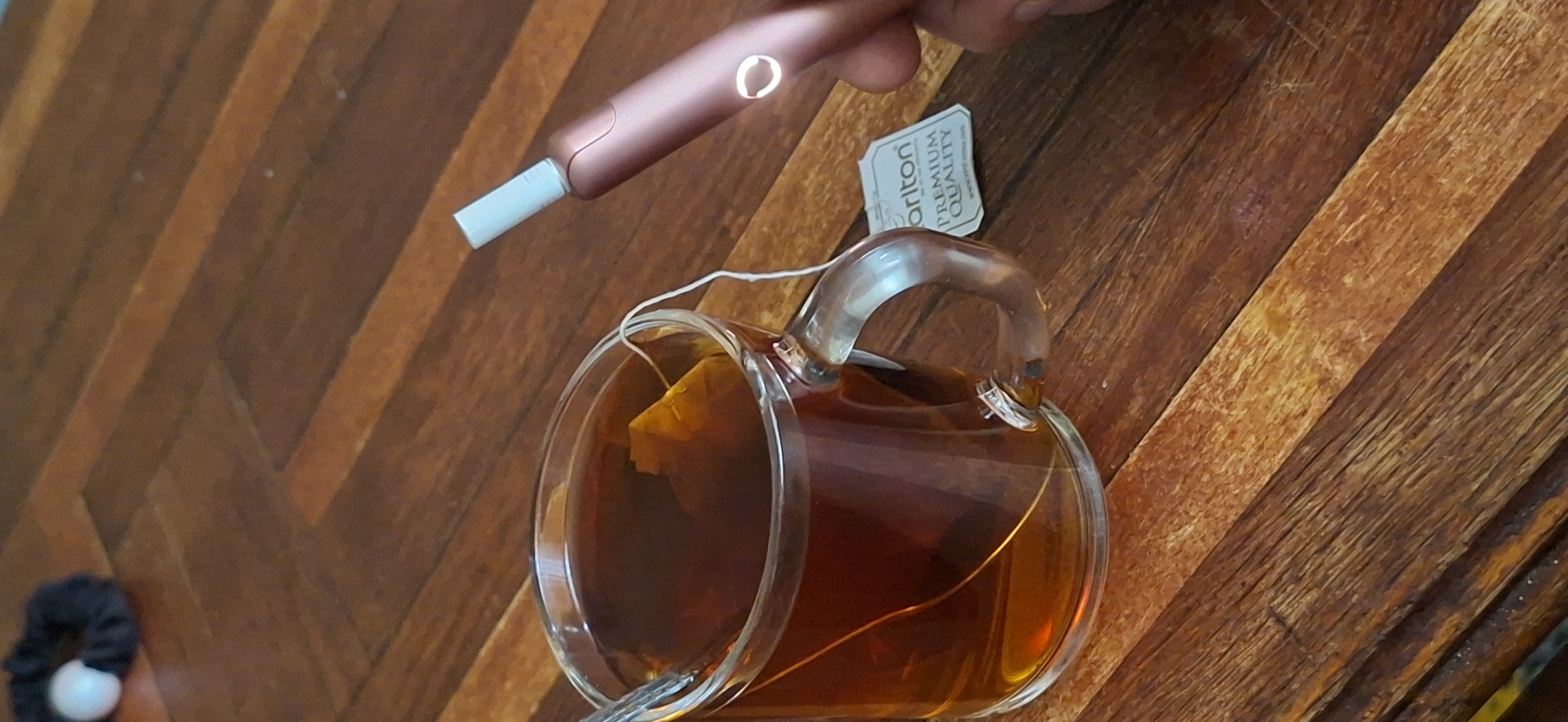 A close-up shot captures a warm beverage in a clear glass mug, accompanied by a smoking device. The mug, filled with dark tea and a tea bag, sits on a wooden surface. A rose-gold colored smoking device, with a white tip, is held by a hand in the right side of the frame, positioned above the mug. A small white tag with the word "Carlton" and "PREMIUM QUALITY" is attached to the mug. In the upper left corner, a black hair tie with a white bead is visible. The background consists of a wooden table with a distinct grain pattern. The image does not contain any elements that would allow for the identification of its specific location in Shakhtarsk, Ukraine, or any other city.