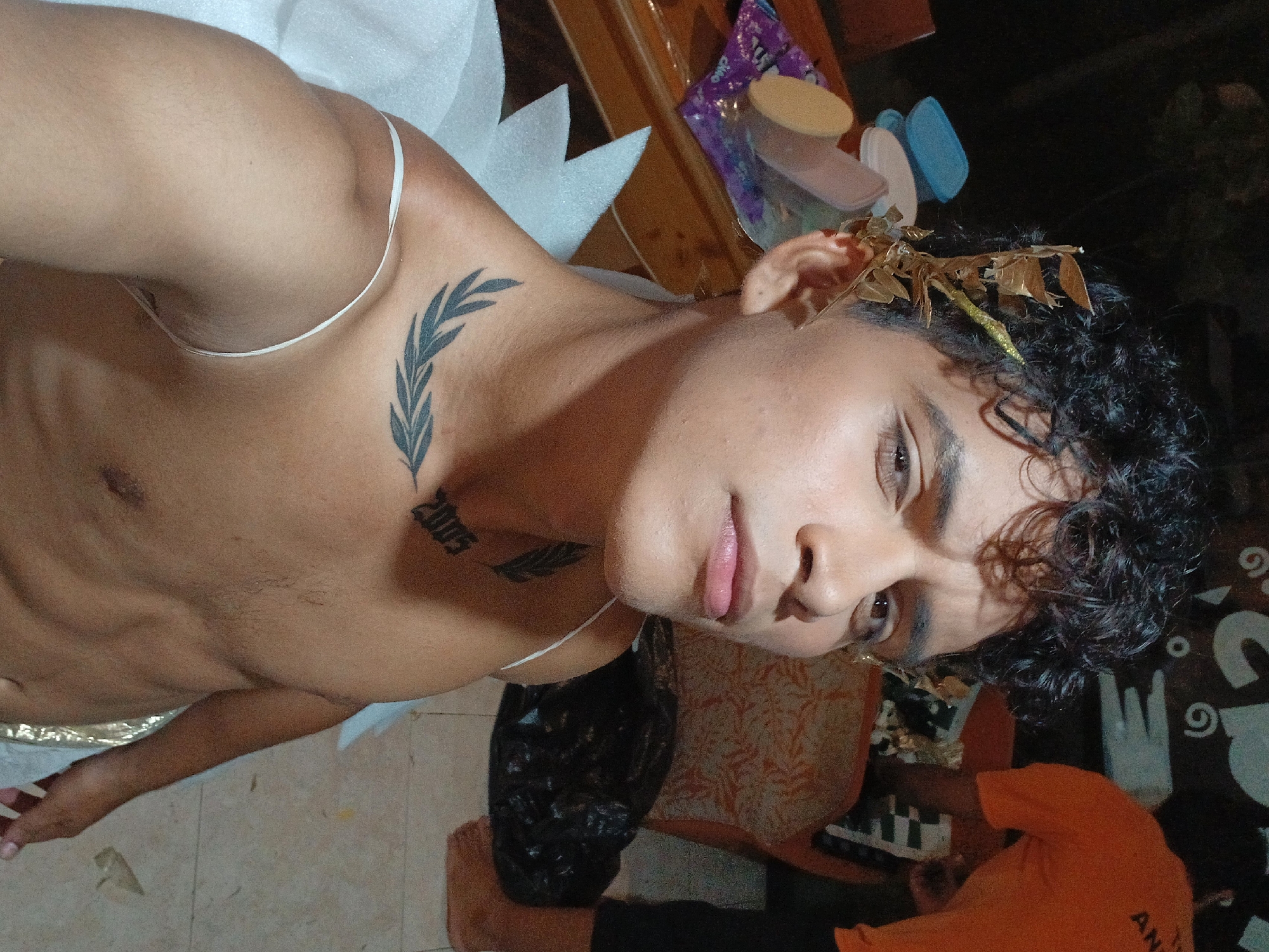 A dimly lit indoor scene shows a light-skinned person with dark curly hair, brown eyes, and bare upper body, adorned with a laurel wreath on their head. Two tattoos are visible: a branch on the left shoulder and the number "2005" on the neck. A thin white strap crosses their left shoulder. The person is posed in a selfie-style shot. The background contains assorted items, including what appear to be craft supplies, and a person in an orange t-shirt partially visible in the lower right corner. There is no information within the image to support the claim that it was taken in Playa El Agua, Venezuela.